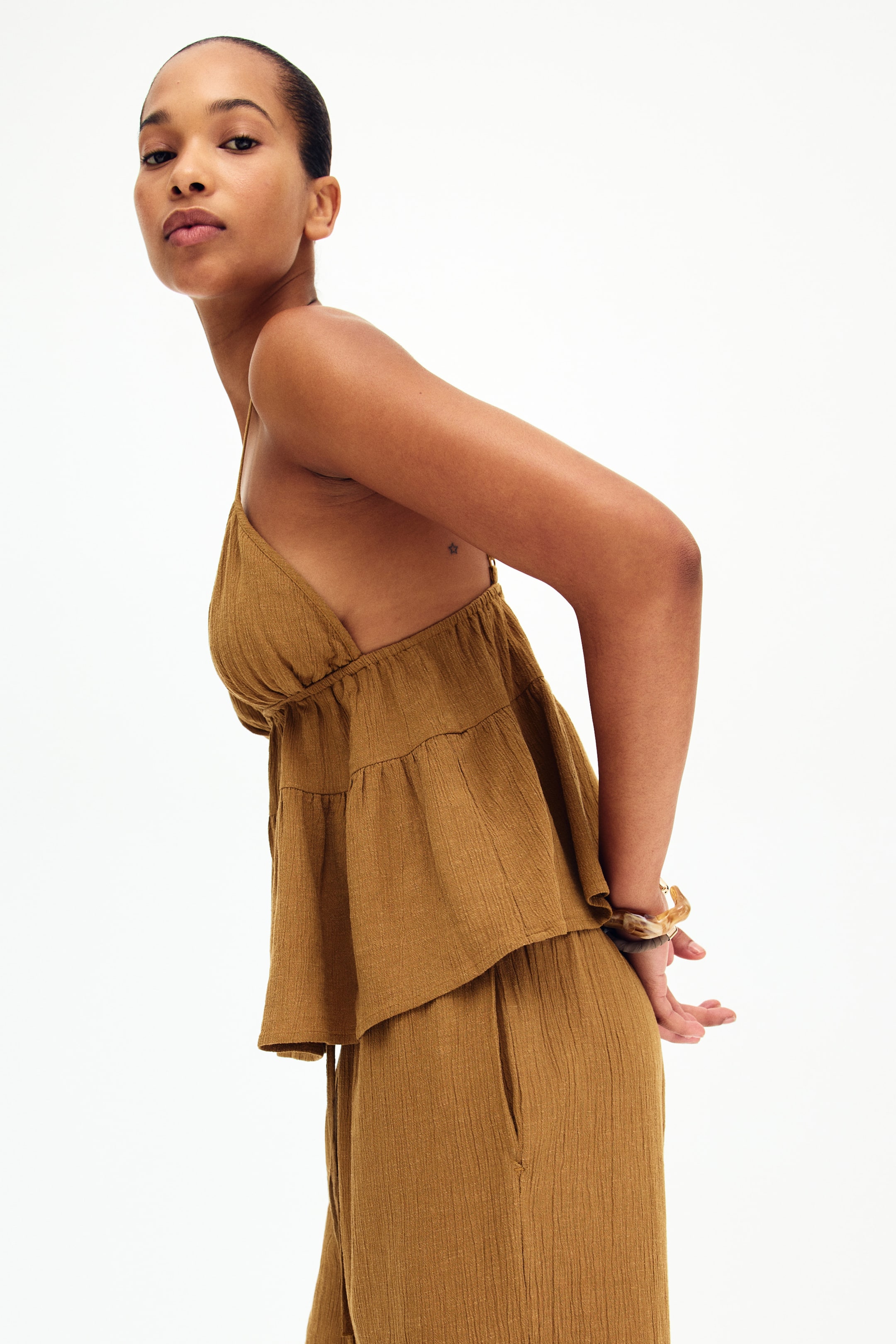 View larger image: An olive brown textured camisole top with thin spaghetti straps, a gathered triangle bodice, and a tiered, flared hem is displayed from a side angle. The matching textured pants, featuring side pockets, perfectly complement the top.