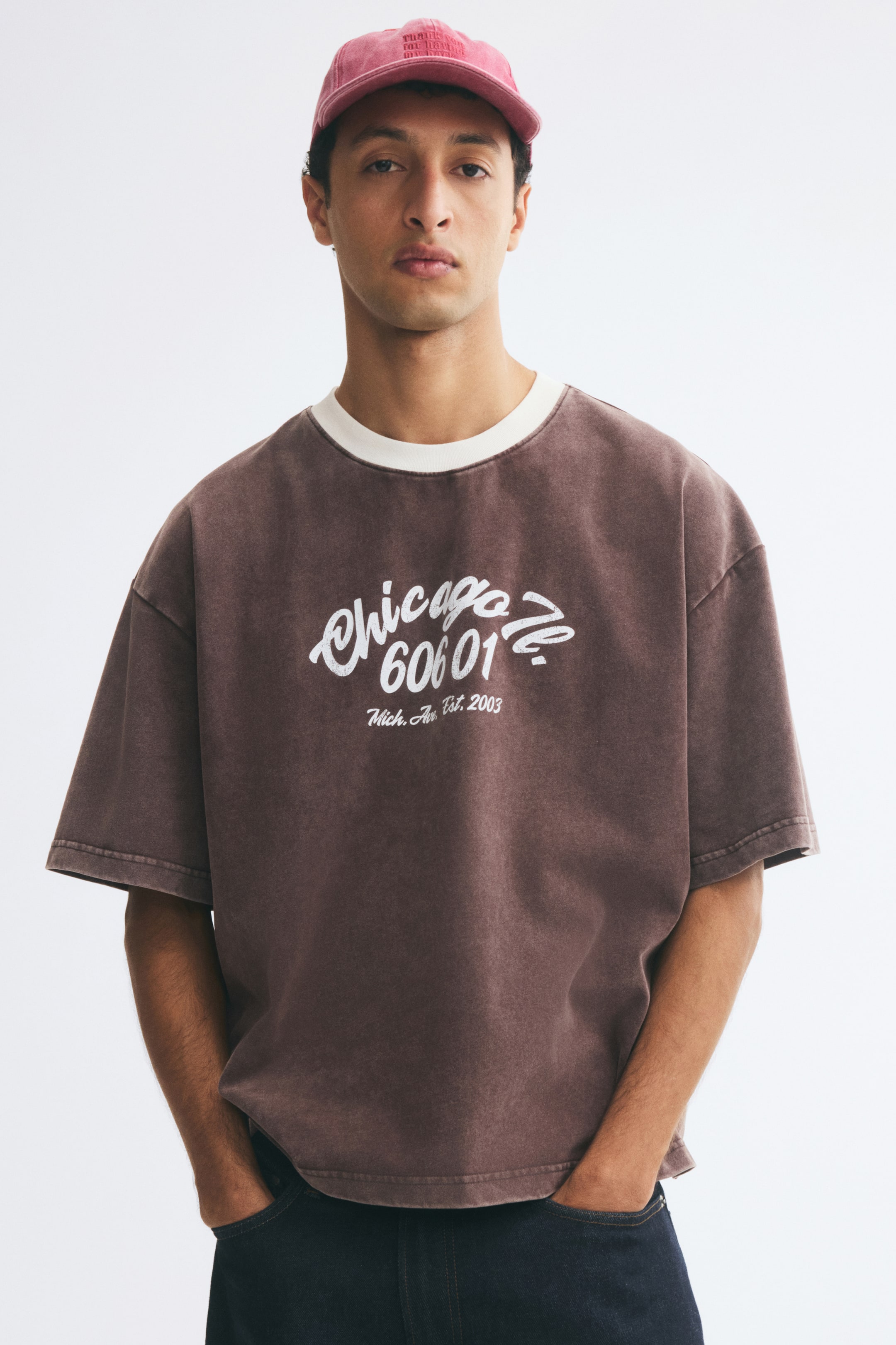 Zobraziť väčší obrázok: Model má na sebe oversized, vyblednuté hnedé tričko z ťažkého bavlneného jersey materiálu. Tričko má na prednej strane bielu grafickú potlač, spustené ramená a kontrastný biely rebrovaný výstrih a je skombinované s tmavými džínsami a vyblednutou červenou šiltovkou.