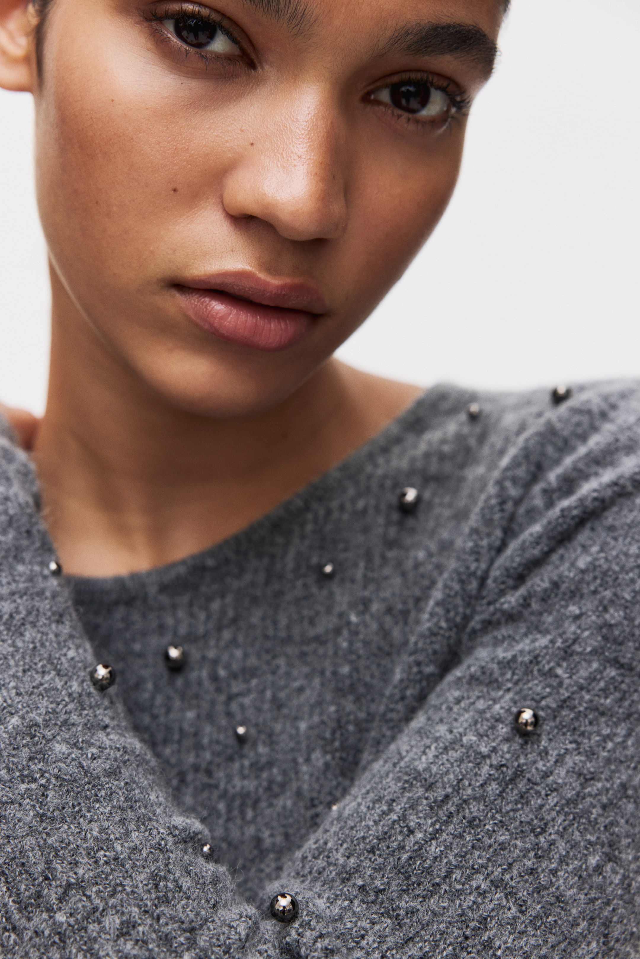 拡大画像を表示: グレーのふわふわしたニットの衣服が展示されています。小さなダークメタリックのスタッズが表面に散りばめられています。衣服はクロスオーバーのVネックラインを形成し、上胸部と肩の周りにスムーズにドレープしています。