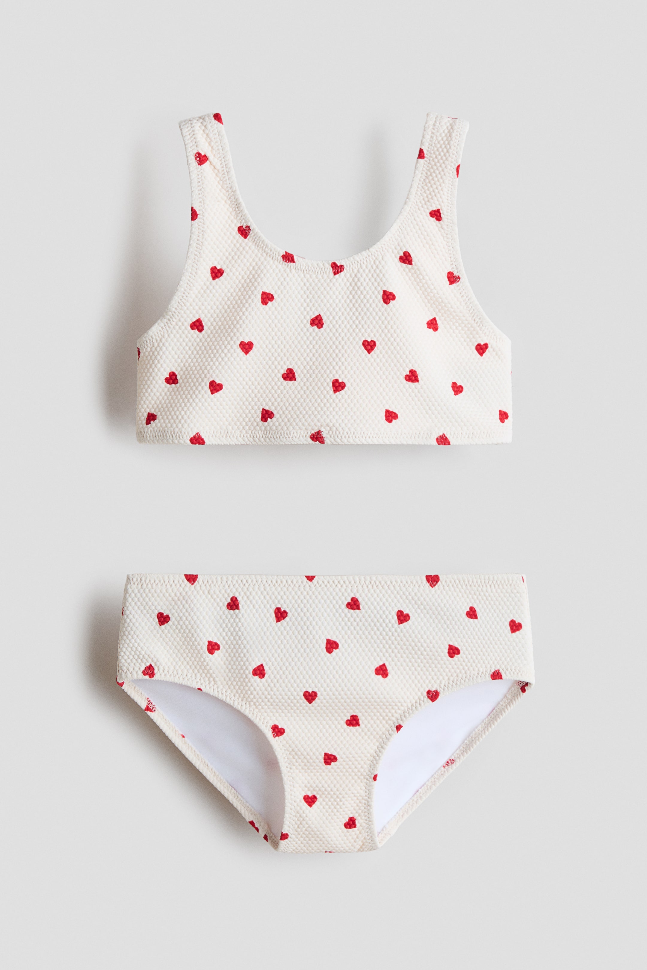 View larger image: A two-piece swimsuit is featuring a cream-colored, textured fabric with small red hearts scattered throughout, presented FRONT. The top is a sleeveless, scoop-neck crop top, and the matching bottom is a standard brief showing white lining at the leg openings.