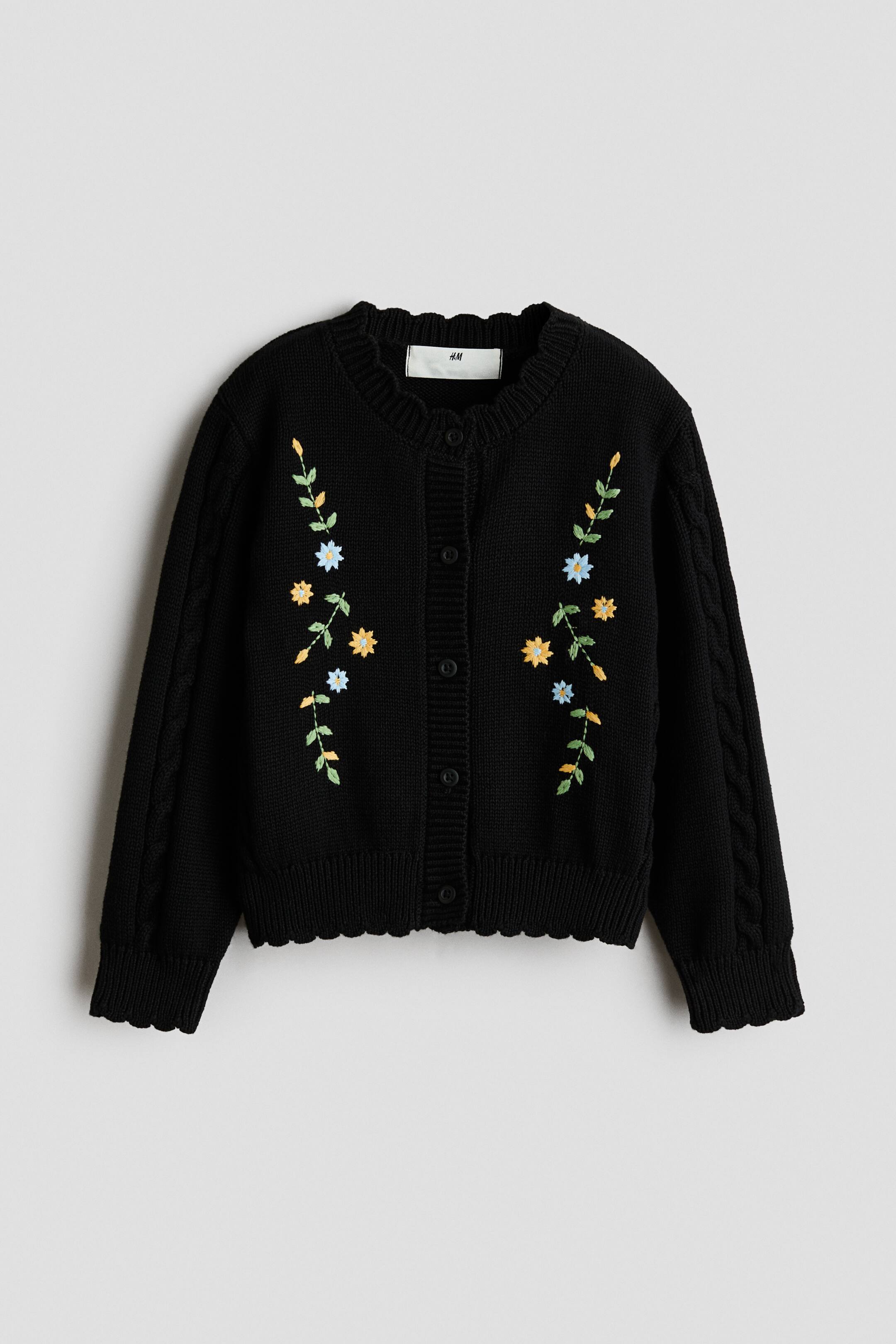 View larger image: A black knit cardigan is shown from the front, featuring colorful embroidered floral vines on either side of the center button closure. The cardigan has a round neckline, dropped shoulders with cable-knit sleeves, and scalloped, ribbed trims along the collar, cuffs, and hem.