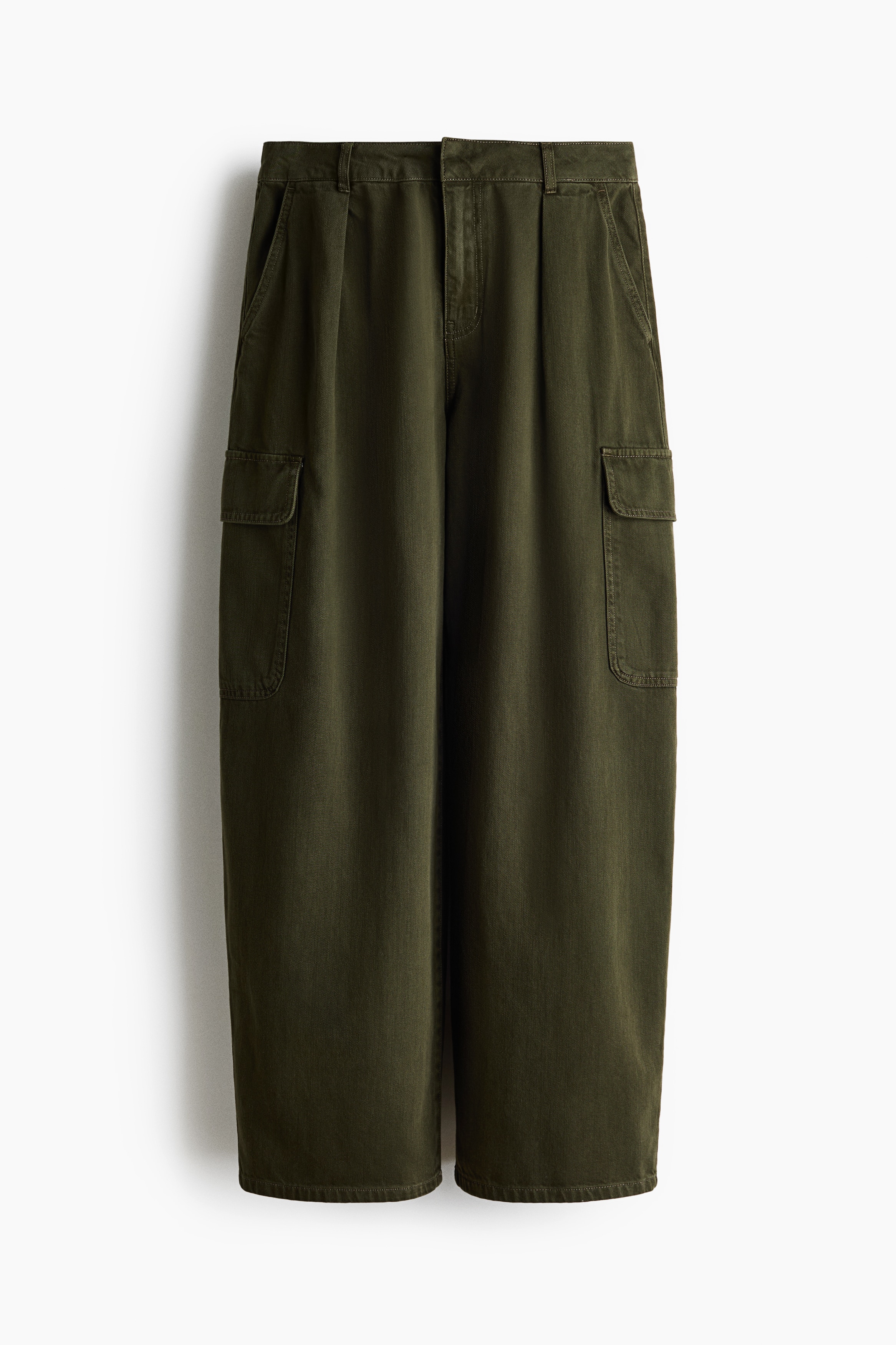 Olive green denim jeans featuring curved, voluminous legs, front pleats, and side patch pockets with flaps are being worn by the model. The loose-fitting, regular-waist jeans are styled with a cream-colored turtleneck sweater and a dark brown padded jacket.