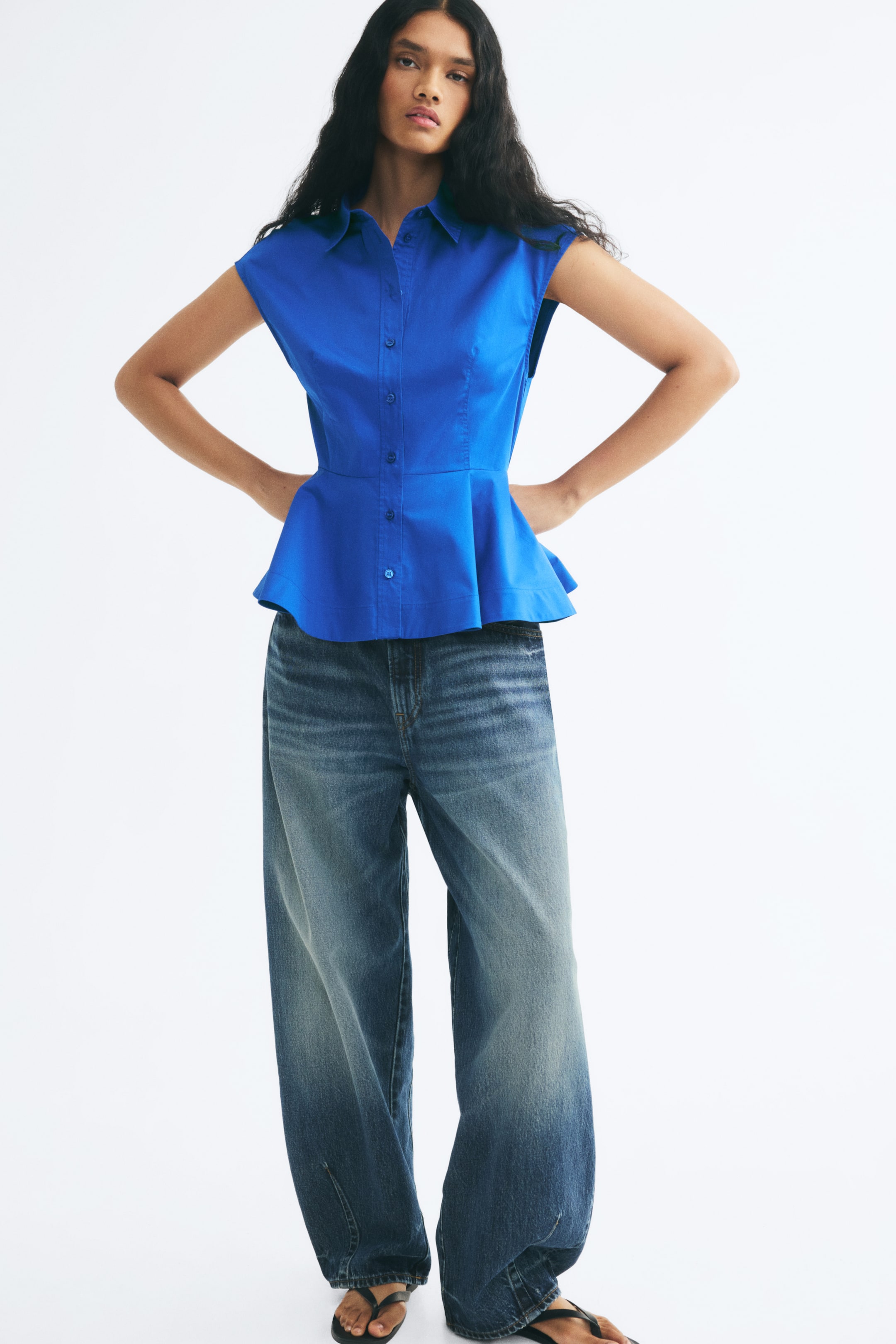 View larger image: A bright blue sleeveless top featuring a collar, button-down front, and flared peplum hem is worn by the model. The top is styled with wide-leg blue denim jeans.
