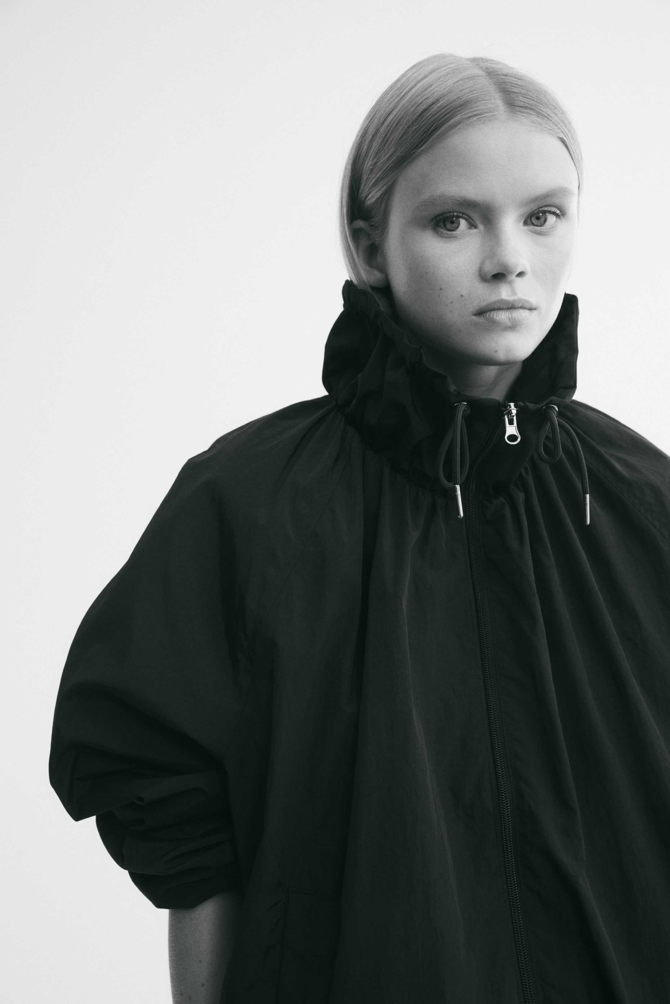 Ver imagem maior: O modelo está a usar uma jaqueta de nylon preta, oversized, com gola alta franzida tipo funil, cordão ajustável e fecho frontal de correr. A jaqueta tem mangas raglan, criando uma silhueta descontraída e volumosa.