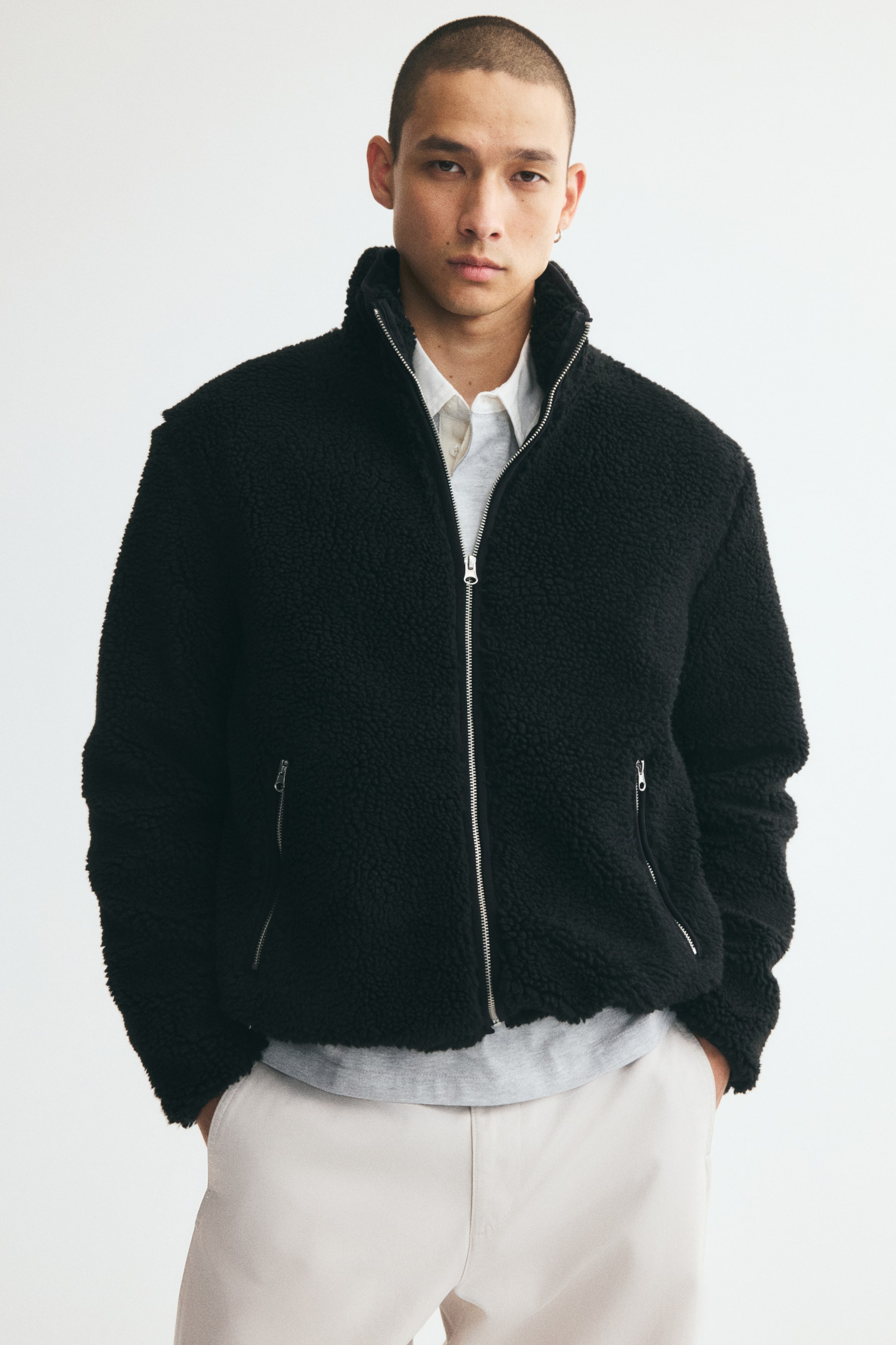 View larger image: A black teddy fleece jacket featuring a stand collar, a front zip closure, and diagonal zippered pockets is worn by the model. The jacket is layered over a white collared shirt and a grey top, paired with light-coloured trousers.