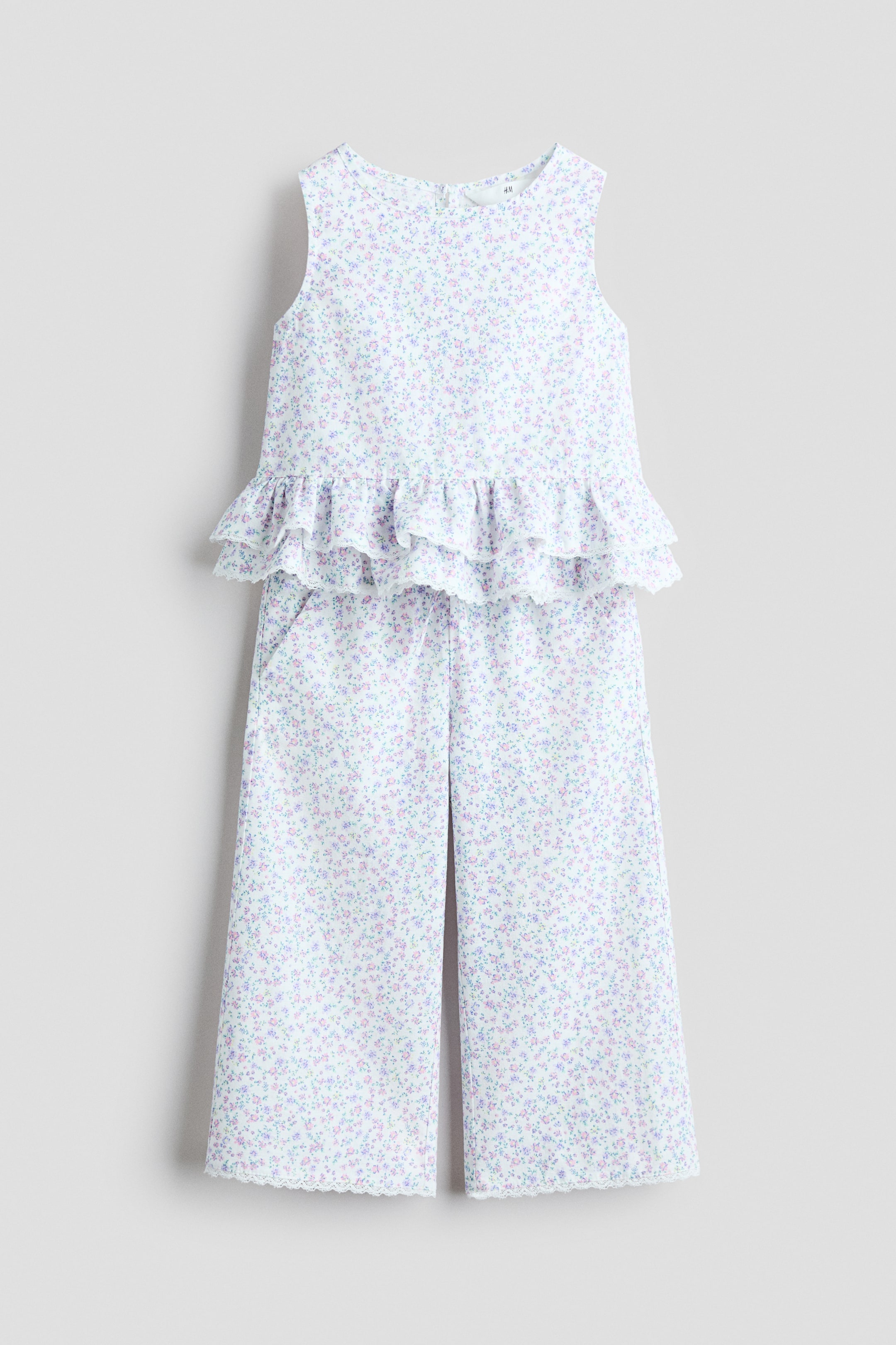 Zobrazit větší obrázek: Přední pohled ukazuje soupravu bez rukávů, která se skládá z topu a širokých kalhot, obojí z bílého materiálu s jemným květinovým potiskem v odstínech fialové, růžové a zelené. Top má kulatý výstřih a stupňovitý volánový lem zdobený krajkovými lemy, zatímco kalhoty mají odpovídající krajkové lemy na širokých nohavicích.