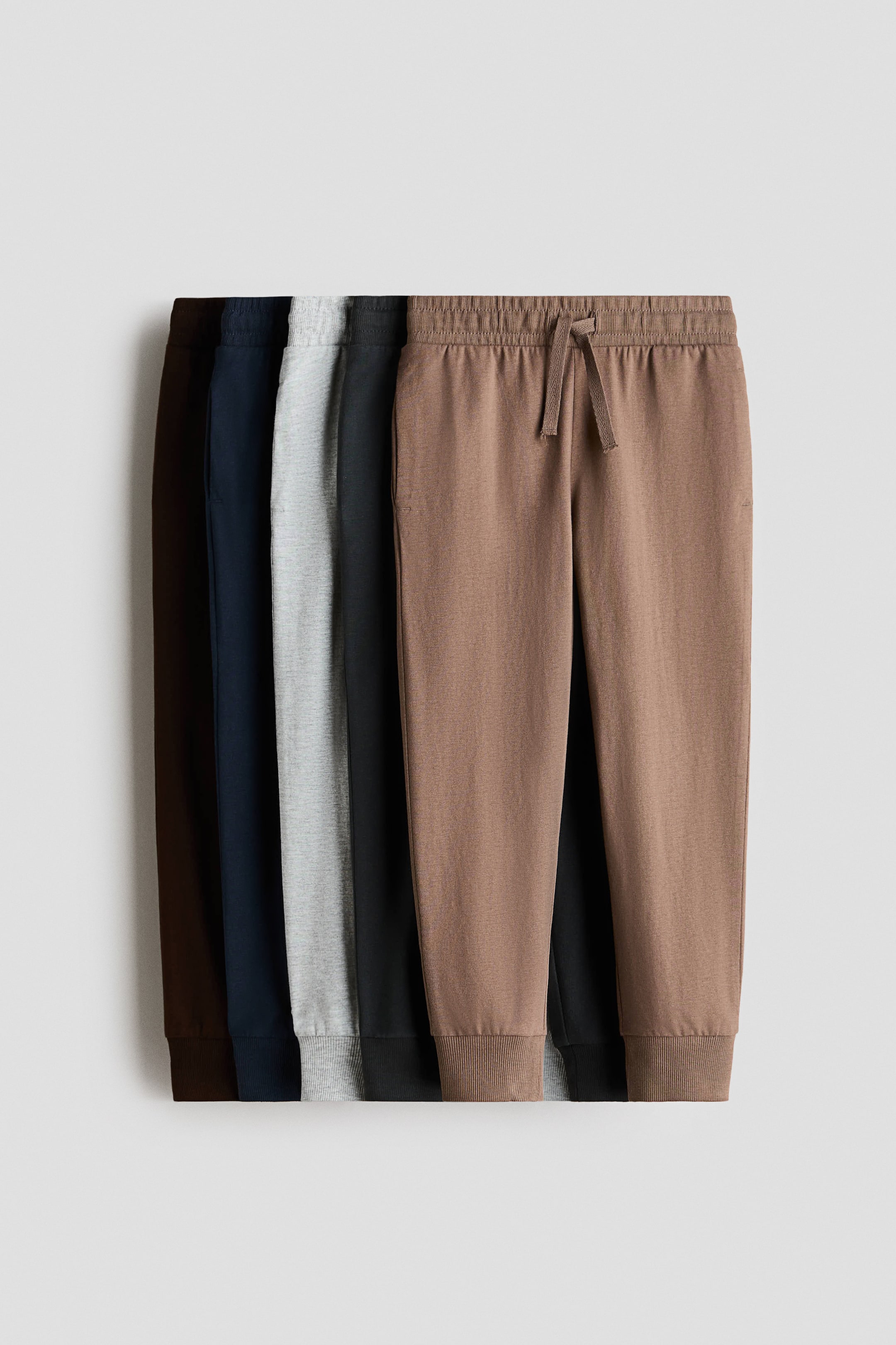 View larger image: Multiple pairs of joggers in soft cotton jersey are being showcased from the front, with the foremost light brown pair featuring an elasticated drawstring waist, discreet side pockets, and ribbed hems. Additional pairs in dark brown, navy, light grey, and black are partially visible behind it.