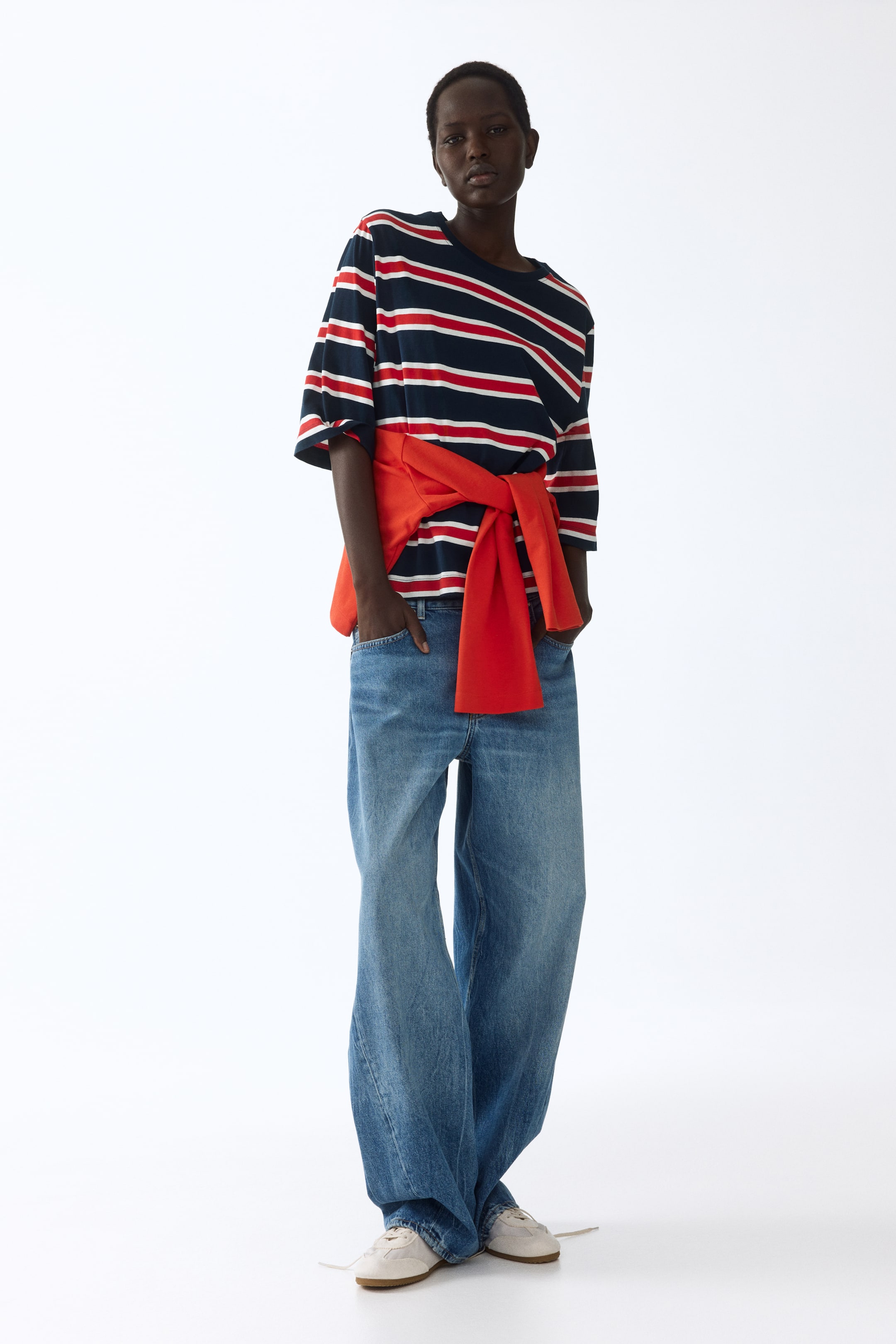 View larger image: This ensemble showcases a navy blue, red, and white striped t-shirt with a crew neck and elbow-length sleeves, paired with light wash, wide-leg denim jeans. A vibrant red sweatshirt is tied around the waist, and light-colored sneakers complete the look.