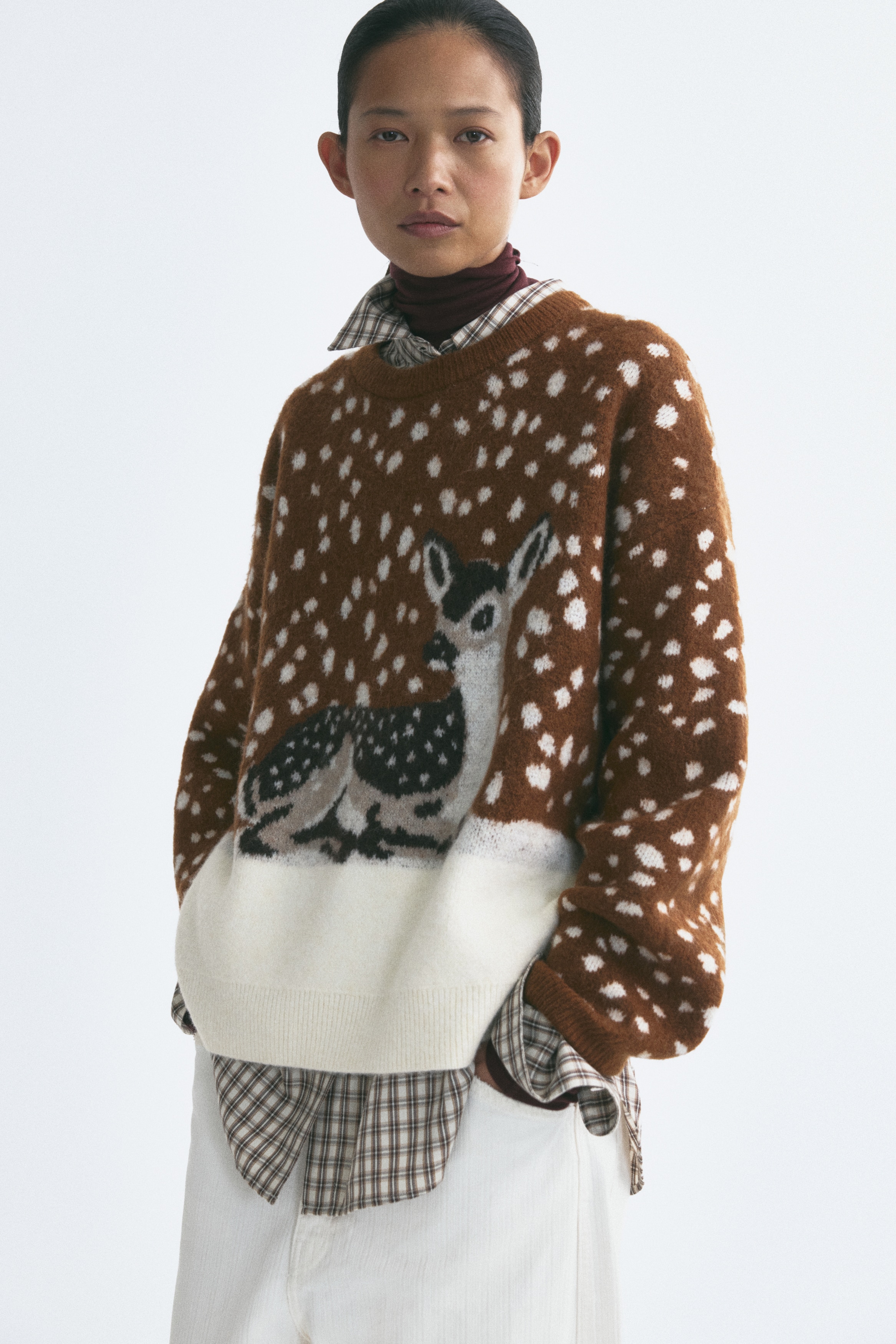 A brown long-sleeved knit sweater is shown from the front, showcasing an all-over white speckled pattern. The centre features a graphic of a spotted deer resting on a solid white lower panel, with ribbed detailing trimming the crew neck, cuffs, and hem.