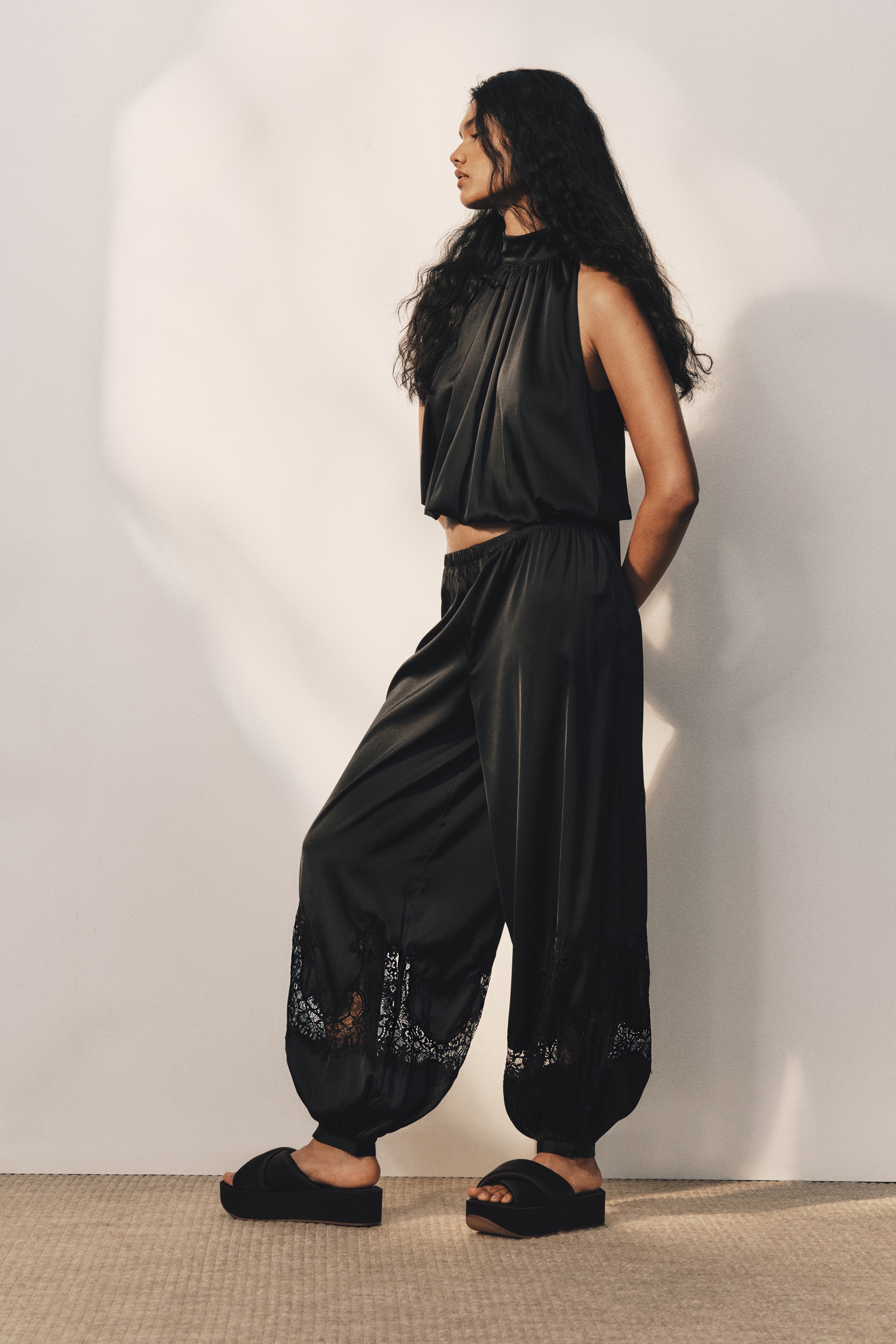 A front view showing black, loose-fit trousers in softly draping satin featuring an elasticated waistband and voluminous balloon legs. The legs are detailed with intricate black lace insets above the ankles, tapering into gathered cuffs with small covered buttons.