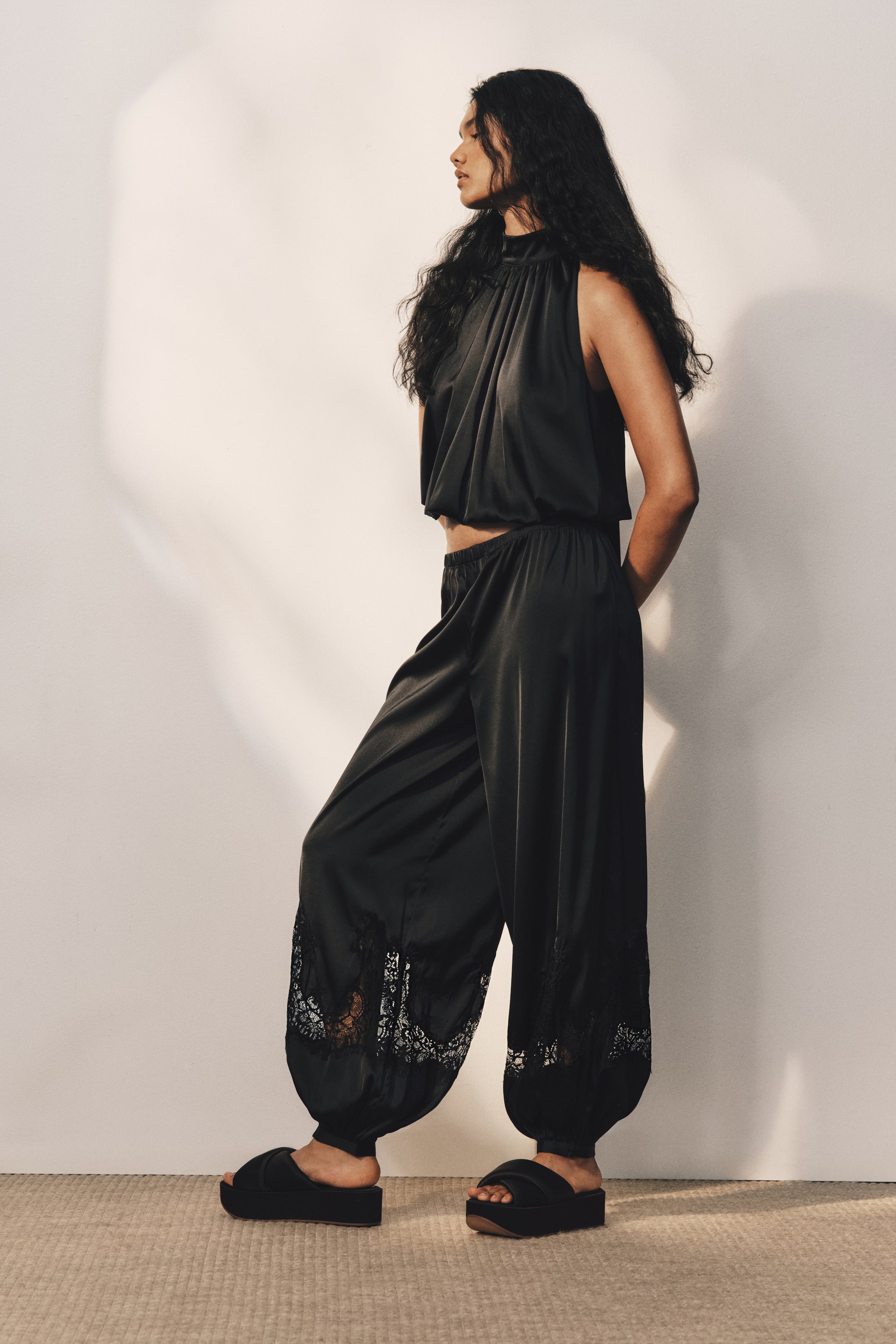 View larger image: Black loose-fit satin pants with an elasticated waist, balloon legs, and sheer lace inserts near the ankles are worn by the model, who is standing in profile. The pants are styled with a matching black satin halter top and black platform slide sandals.