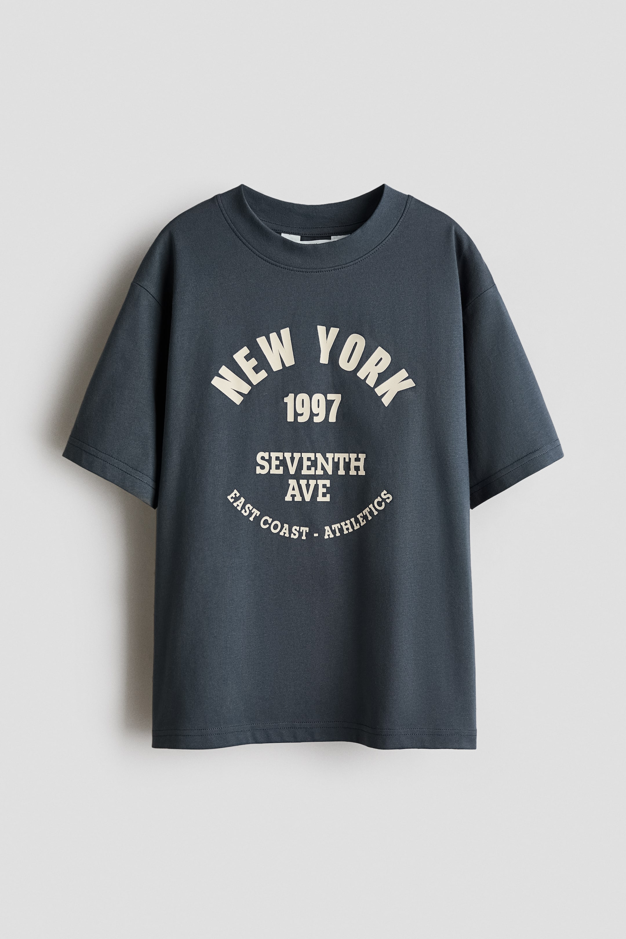 View larger image: A dark grey short-sleeved t-shirt is facing FRONT, featuring a ribbed crew neck and a relaxed fit. The front is displaying a cream-colored graphic print with "NEW YORK" arced at the top, "1997" and "SEVENTH AVE" centered below, and "EAST COAST - ATHLETICS" arced at the bottom.
