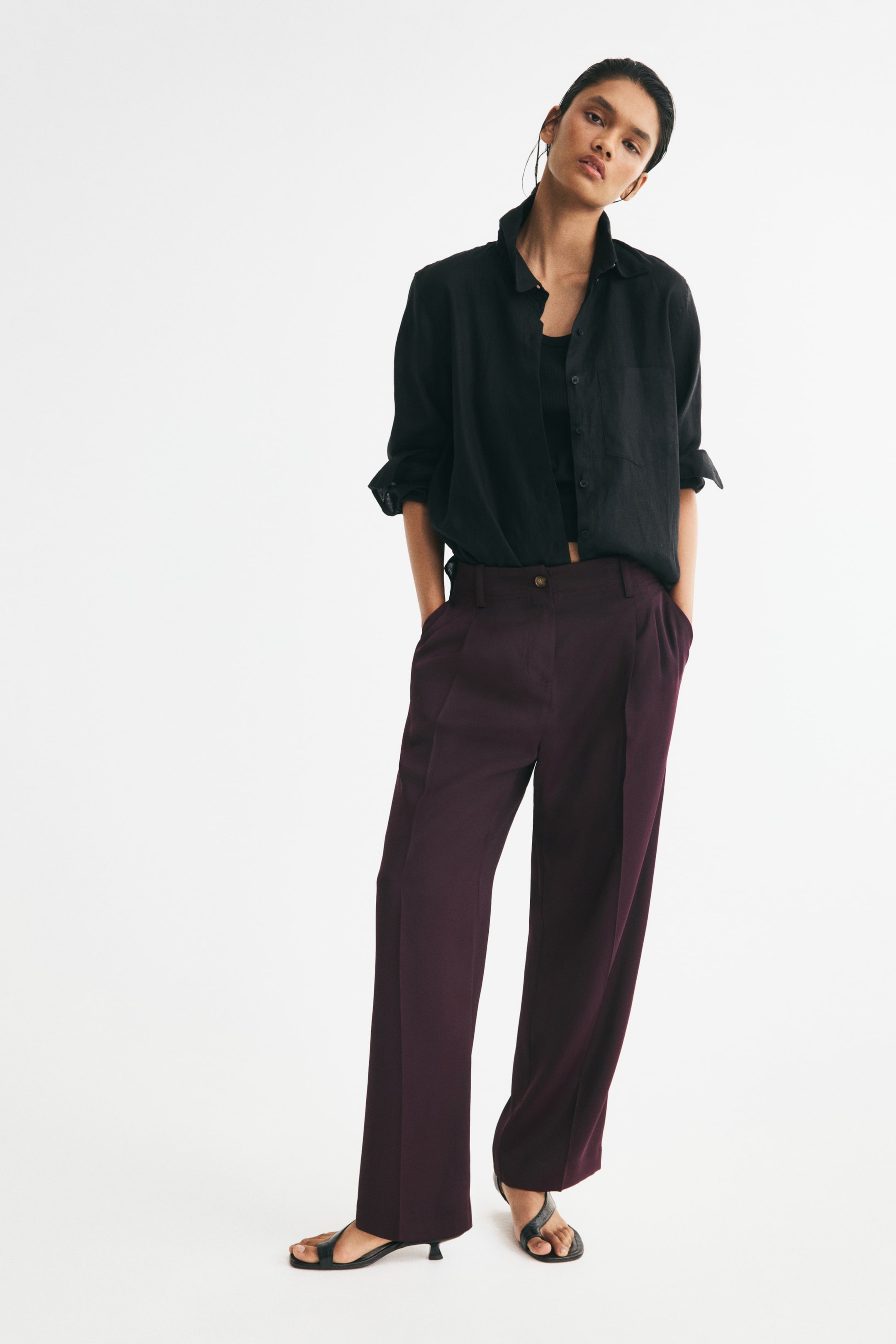 View larger image: Relaxed-fit trousers in deep plum draping satin are worn by the model, featuring a visible button closure, barrel legs with pleats at the top, front creases, and diagonal side pockets where the model is placing her hands. The model styles the trousers with a black shirt and black sandals.