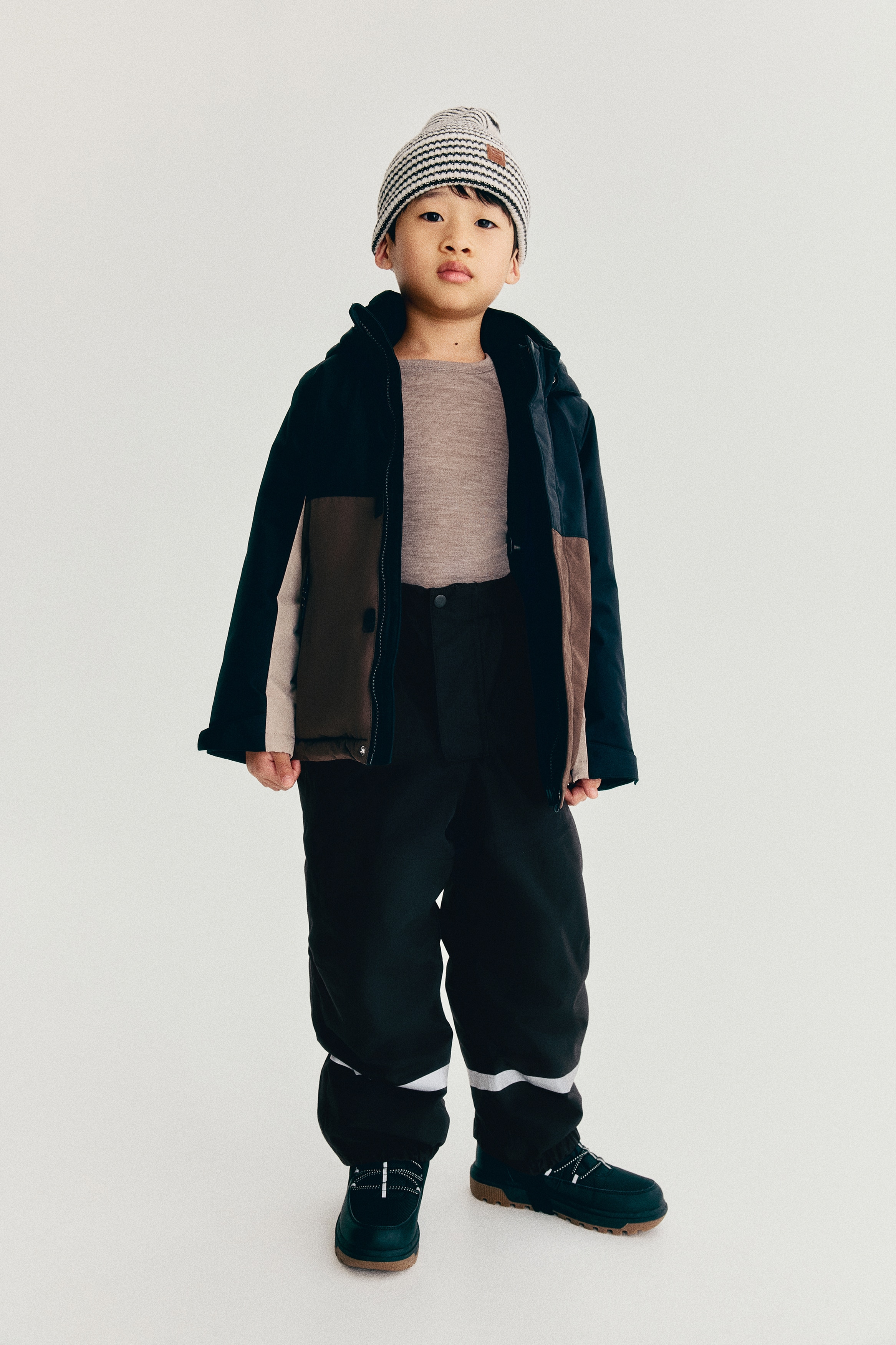 A pair of black childrenâs snow trousers is shown from the front, featuring adjustable elastic braces and an elasticated waistband with a snap-button placket. The trousers feature horizontal reflective bands near the lower legs and elasticated ankle cuffs with underfoot stirrup straps.