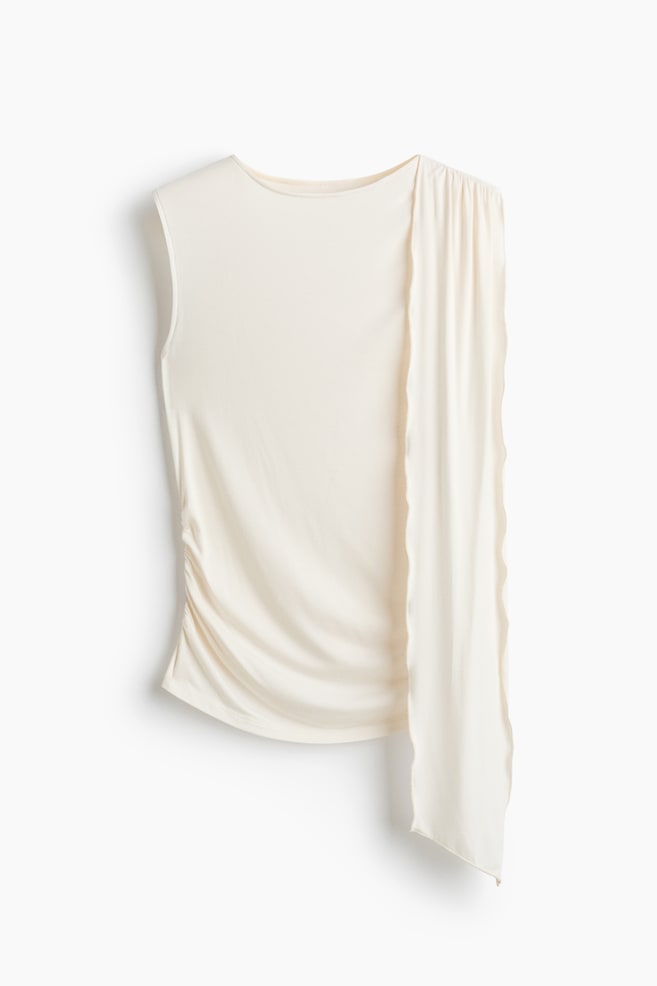 A cream sleeveless top is featuring a boat neck and ruching along its left side. A long, draped panel is extending from the right shoulder, falling past the hem. - 1