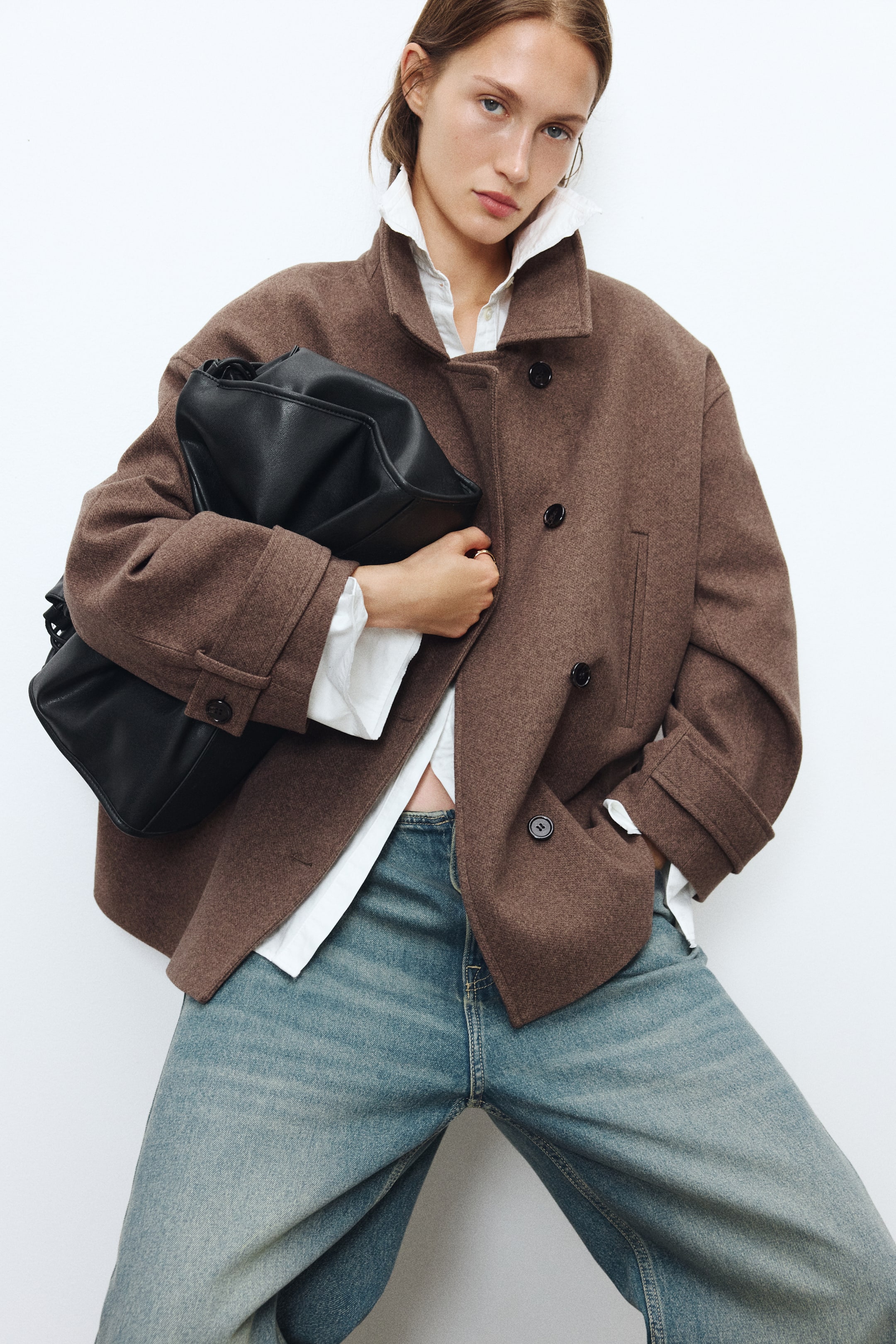 View larger image: A brown felted jacket featuring a funnel collar, dropped shoulders, and buttoned tabs at the cuffs is being worn by the model over a white collared shirt and blue jeans. The jacket is styled with offset front buttons, and the model is carrying a large, slouchy black bag tucked under one arm.