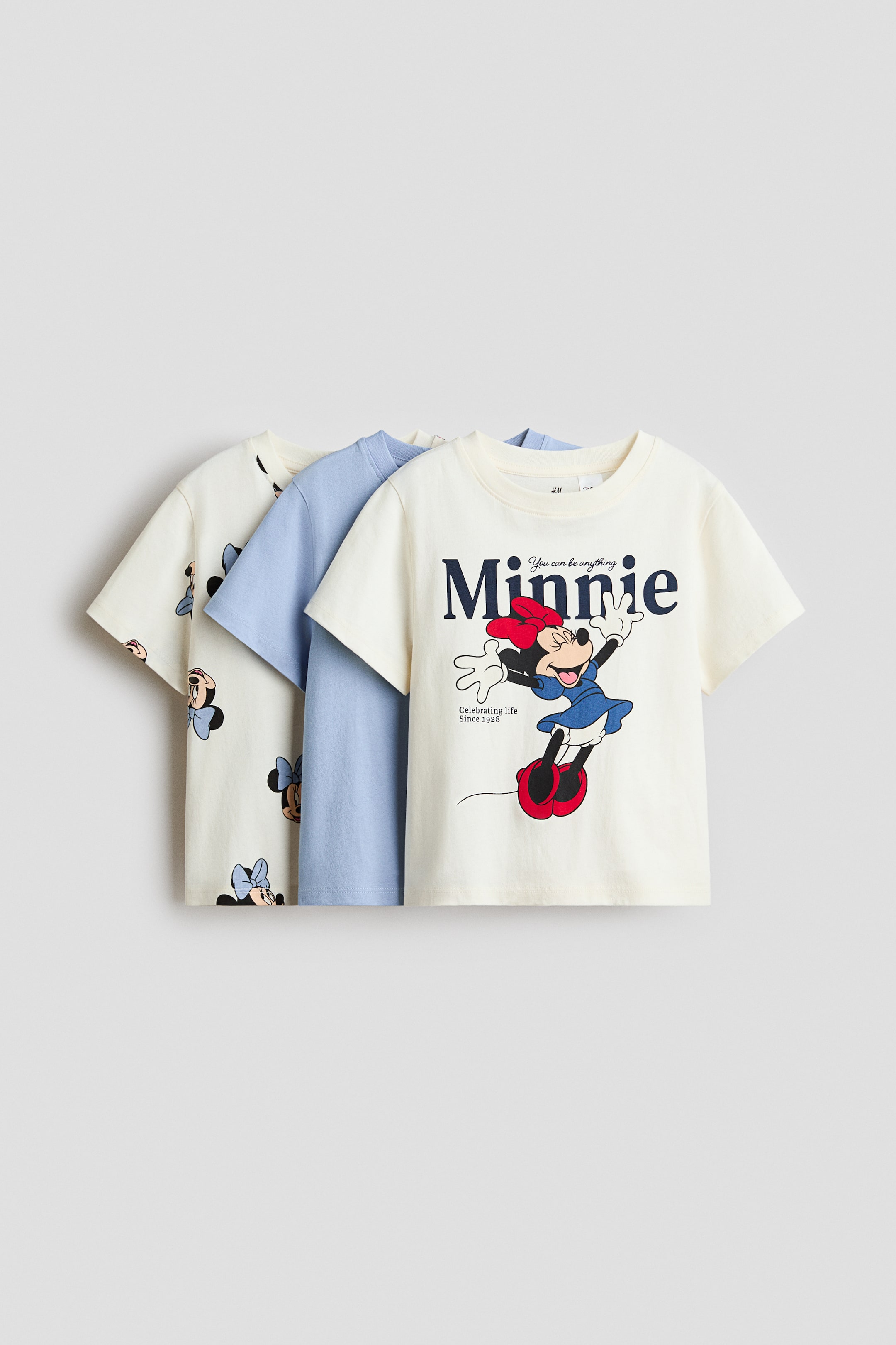 View larger image: Three short-sleeved tops are layered, with the front off-white top featuring a graphic print of Minnie Mouse in a blue dress and red shoes, with "Minnie" text above. A plain light blue top is visible in the middle, and a third off-white top with a repeating Minnie Mouse head pattern is partially showing behind it.