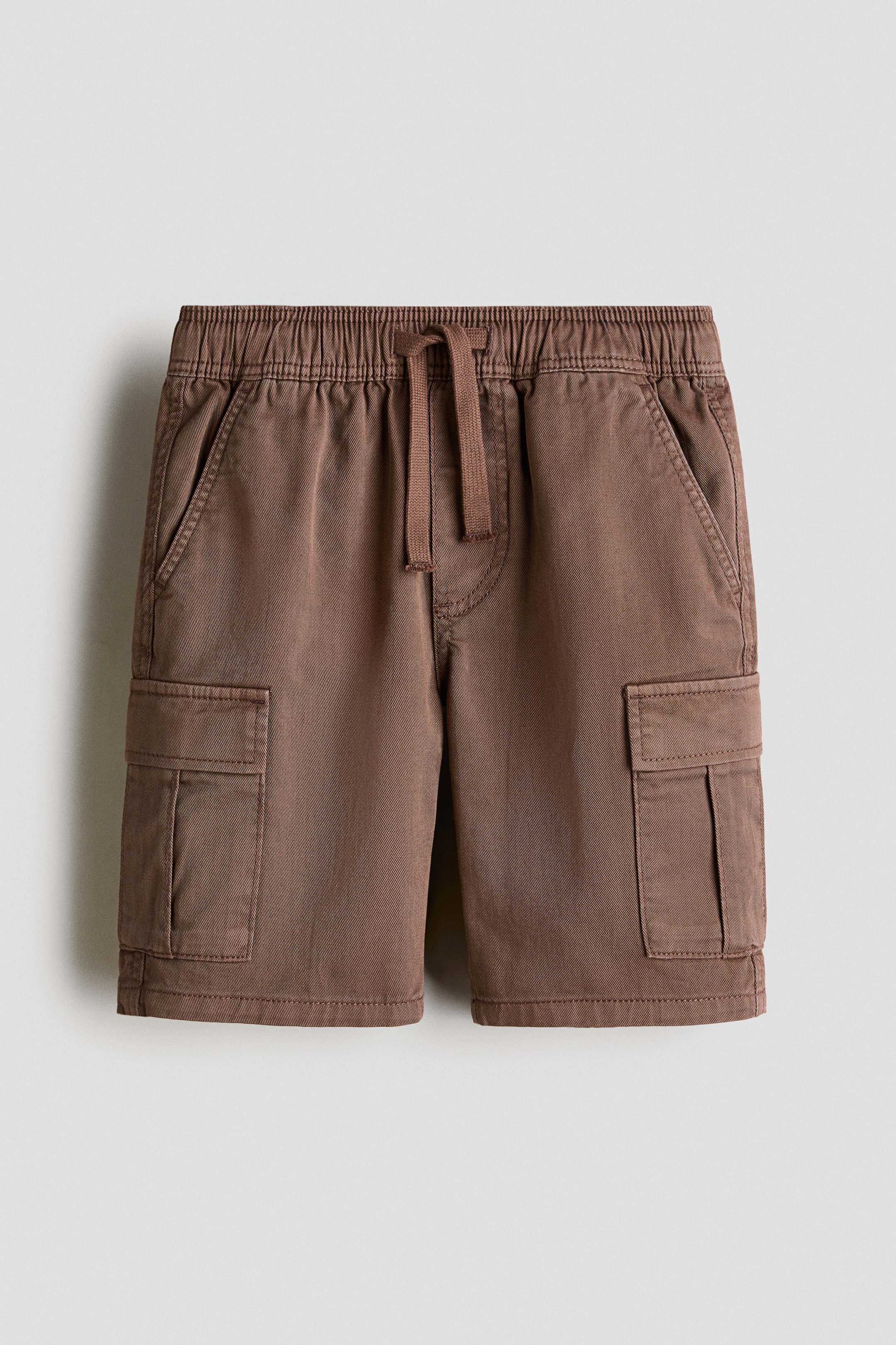 View larger image: The brown cargo shorts are showcased from the front, highlighting an elasticated waistband with a central drawstring tie. Two front slant pockets and two side cargo pockets with flap closures are visible.