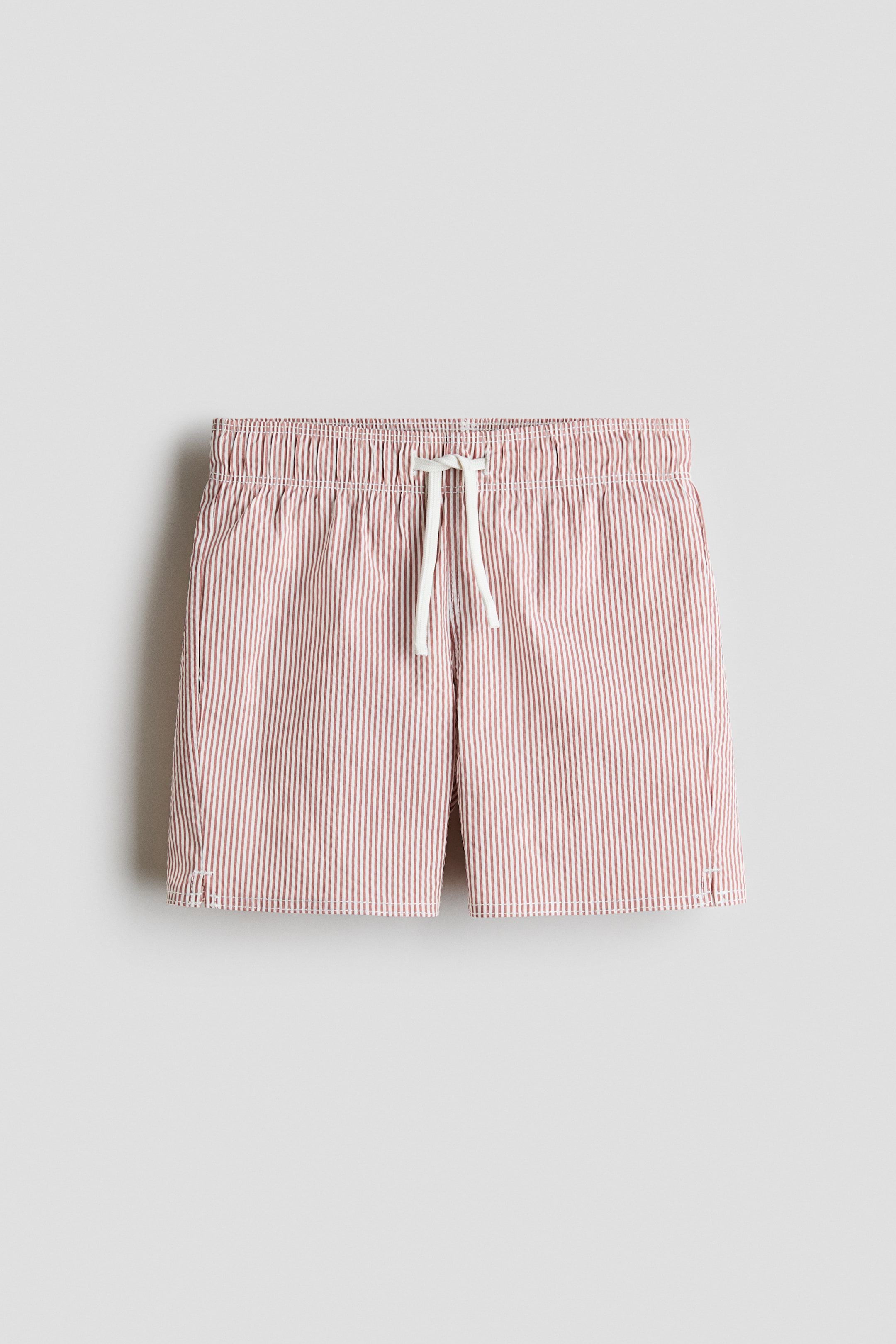 View larger image: H&M Adorables swim shorts are presenting a front view, featuring reddish-brown and white vertical stripes. They are designed with an elasticated, drawstring waist, and small slits are visible on the side hems.