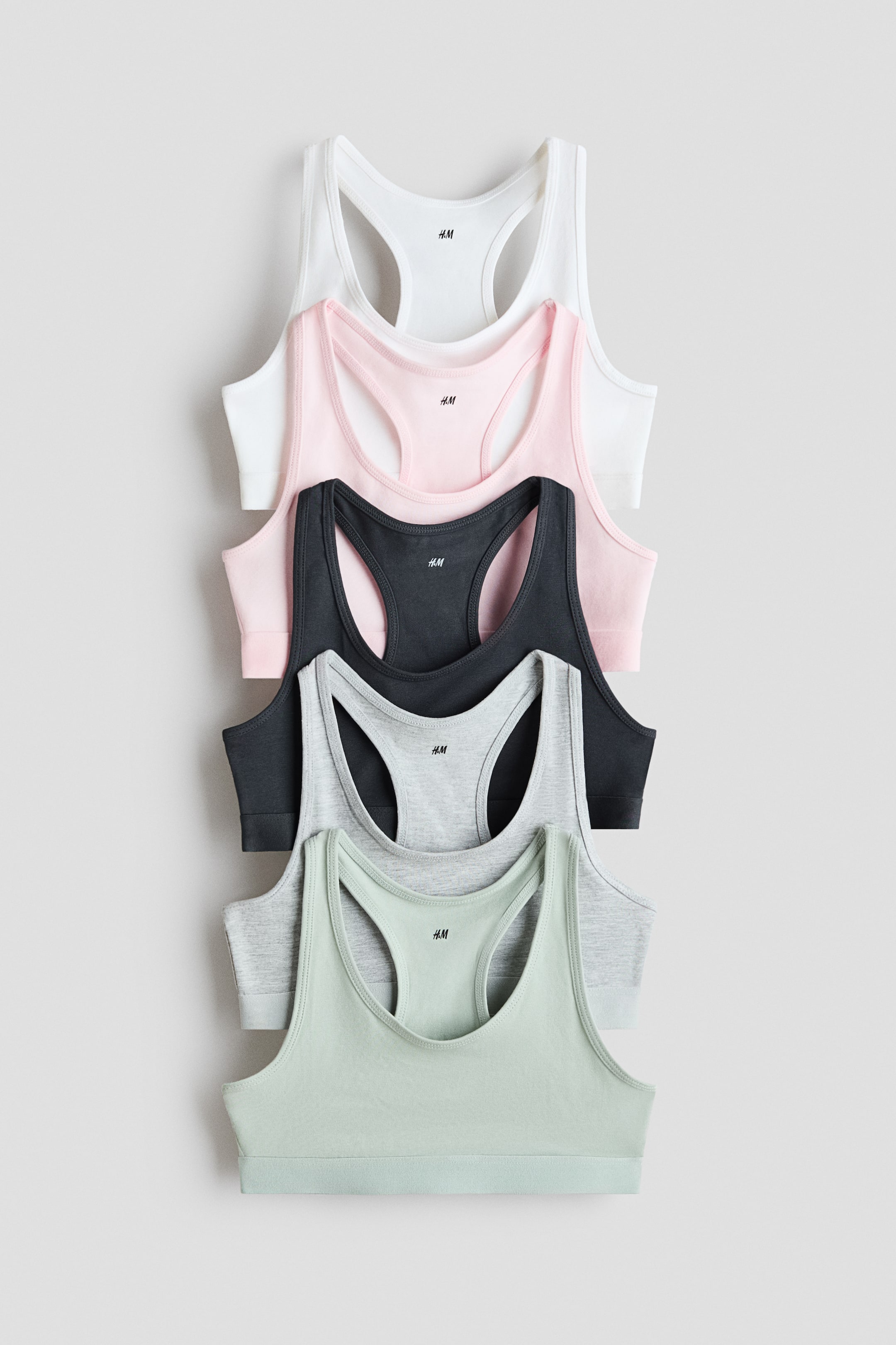 View larger image: Five soft-cup bralettes are stacked, showcasing a white, light pink, dark gray, light gray, and light green garment. Each bralette features a racerback design and a small "H.M" logo on the front center.