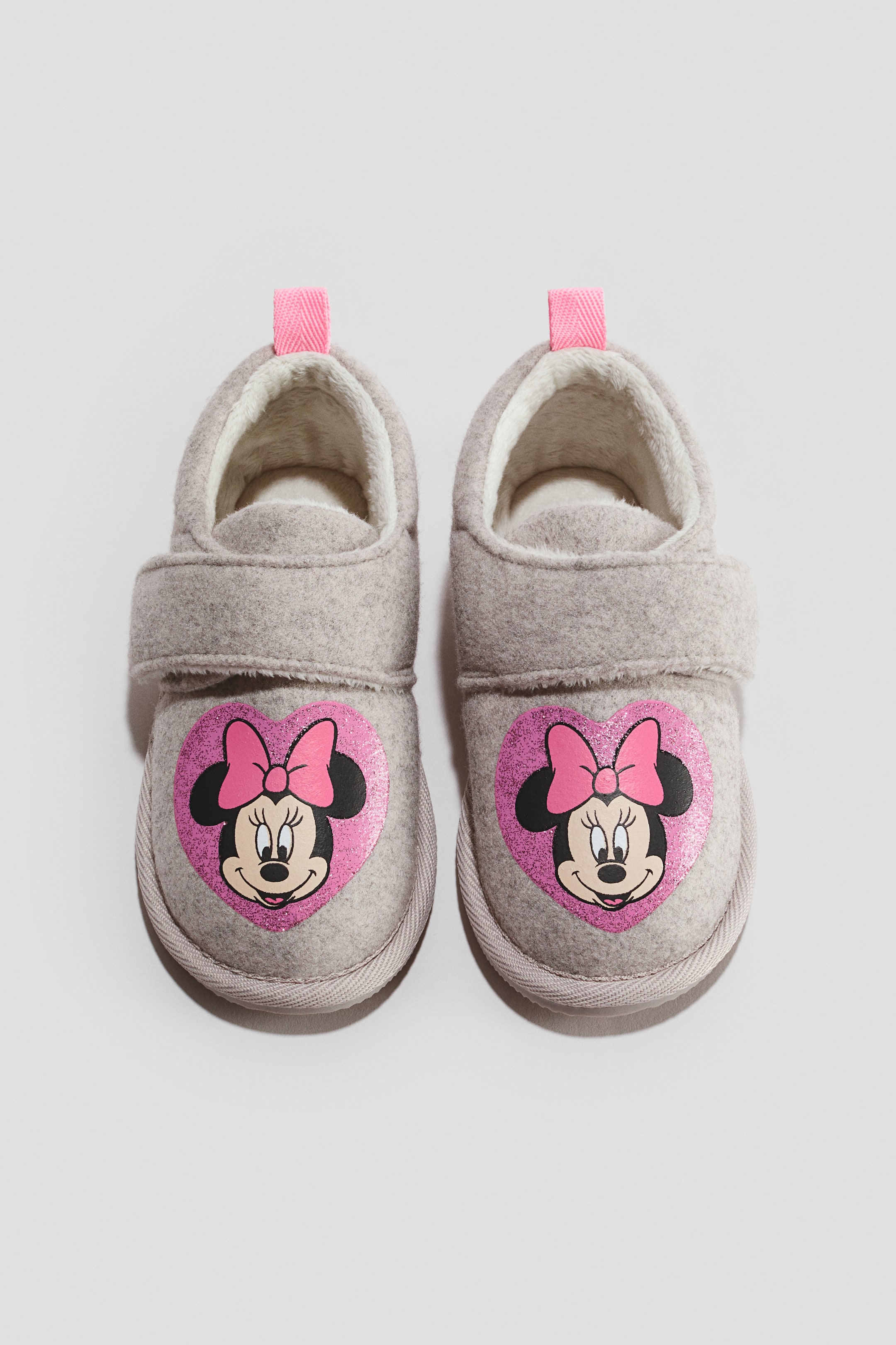 Visa större bild: Ett par ljusgrå barns tofflor i filt visas framifrån, med en enkel kardborreknäppning och ett rosa glittrigt hjärtmotiv med Minnie Mouse på varje tå. Tofflorna visar upp ett mjukt, fluffigt vitt innerfoder och en rosa dragflik i vävband vid hälen.