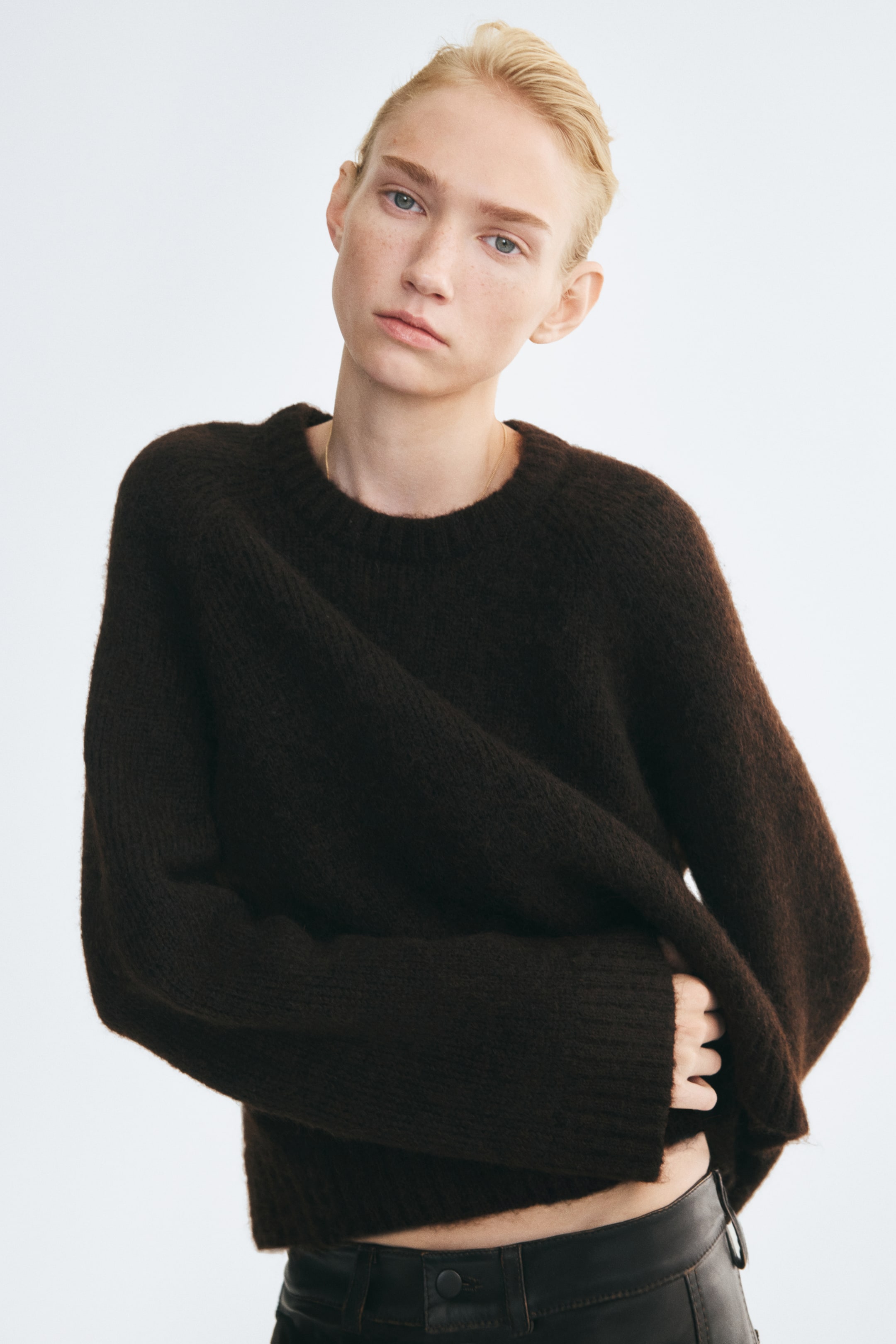Grotere afbeelding bekijken: Een oversized, donkerbruine trui van zachte knit wordt door het model gedragen, met een ronde, geribde halslijn, lange mouwen met geribde boorden en een geribde zoom. Het model houdt met één hand de zijkant van de trui ter hoogte van de taille vast en trekt deze iets samen, waardoor de tailleband van een donkerleren broek zichtbaar wordt.
