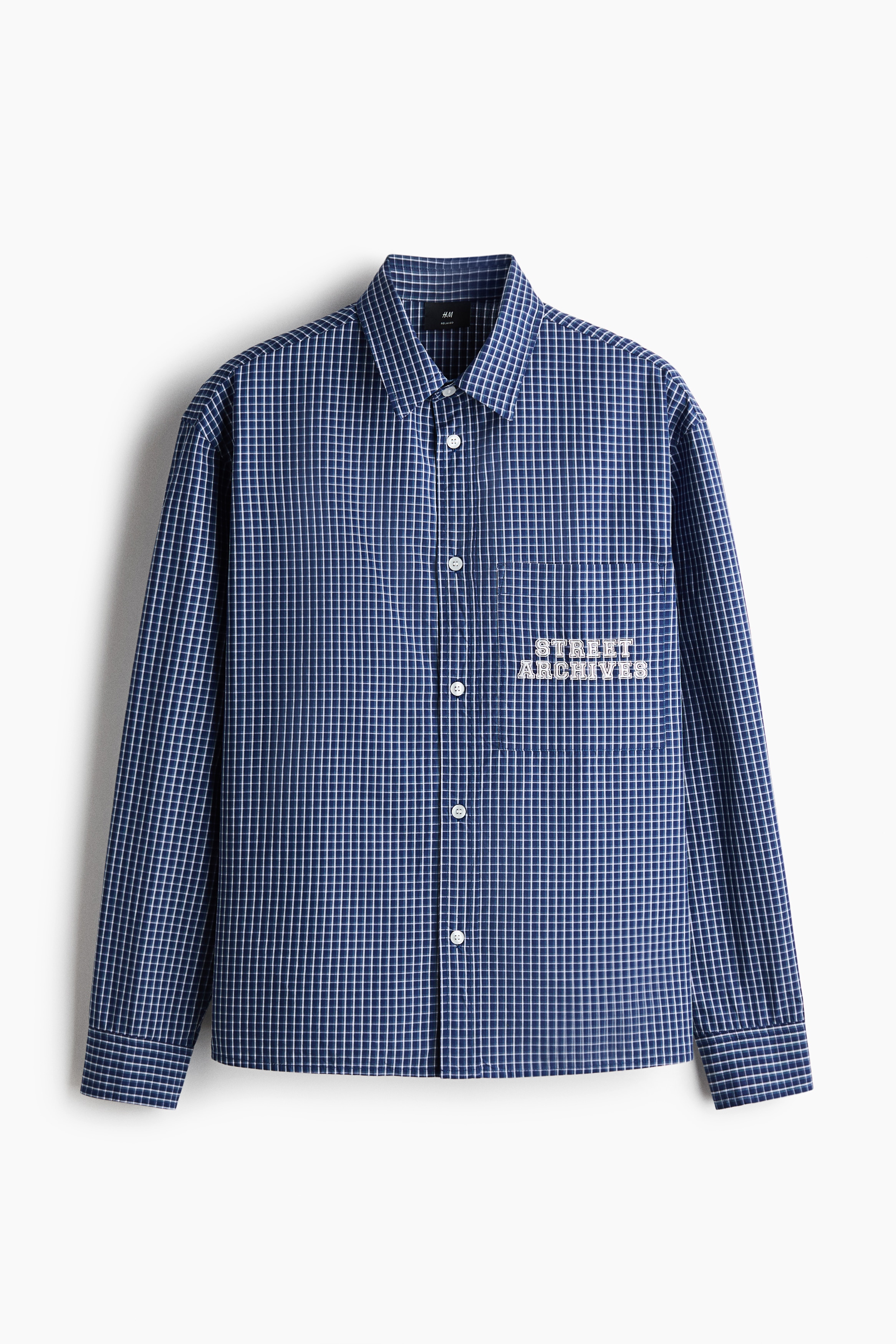 A blue and white checkered cotton poplin shirt is worn open by the model, layered over a cream-coloured knit top and dark blue jeans. The relaxed-fit shirt features a classic collar, dropped shoulders, and a patch pocket on the chest with white printed text.