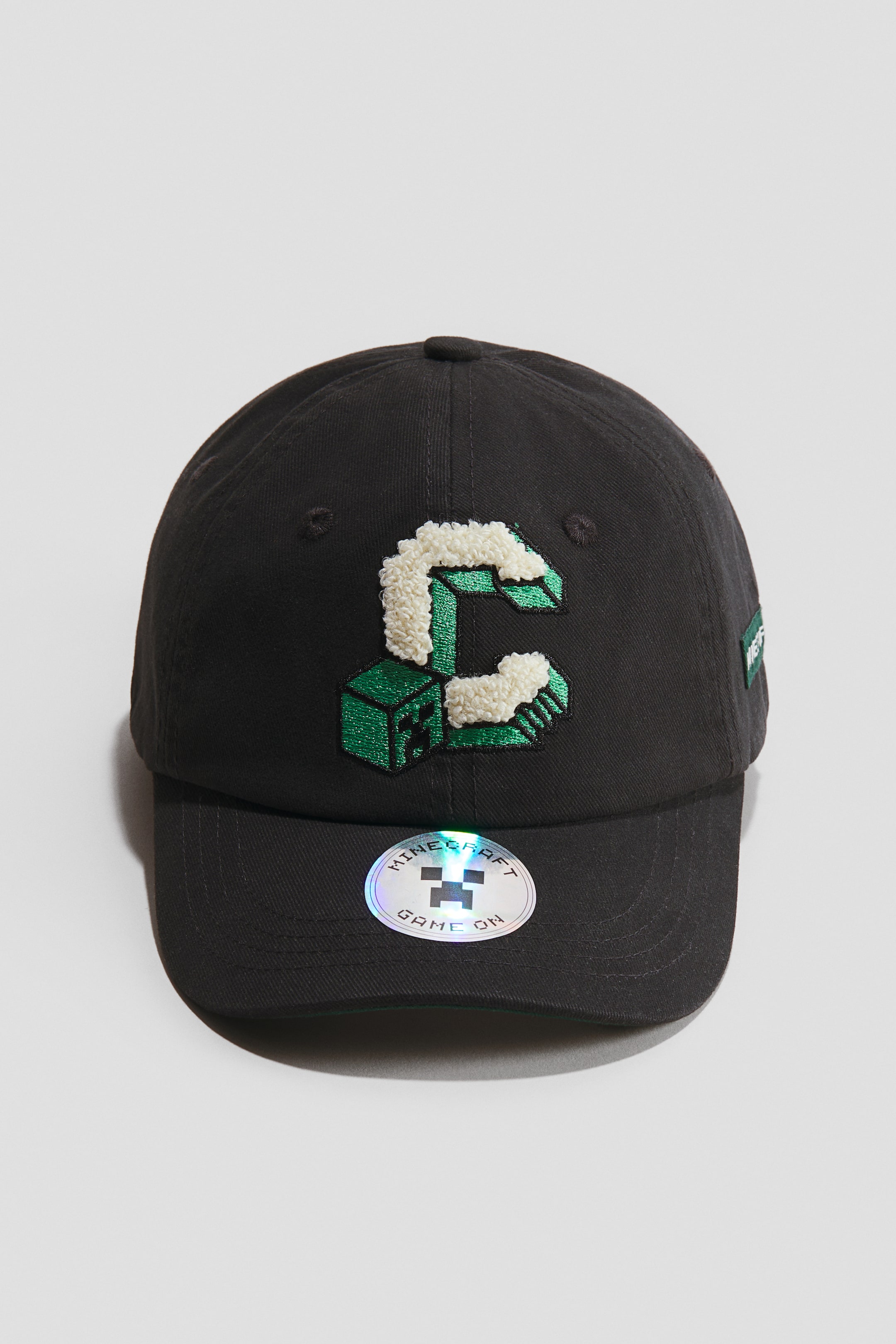 View larger image: A dark grey cap is facing front, showcasing motif details on the crown, including a large embroidered 'C' made of green pixelated blocks and white textured material, with an additional green pixelated block below it. Embroidered eyelets are visible on the top, and a holographic Minecraft sticker is adhering to the brim.