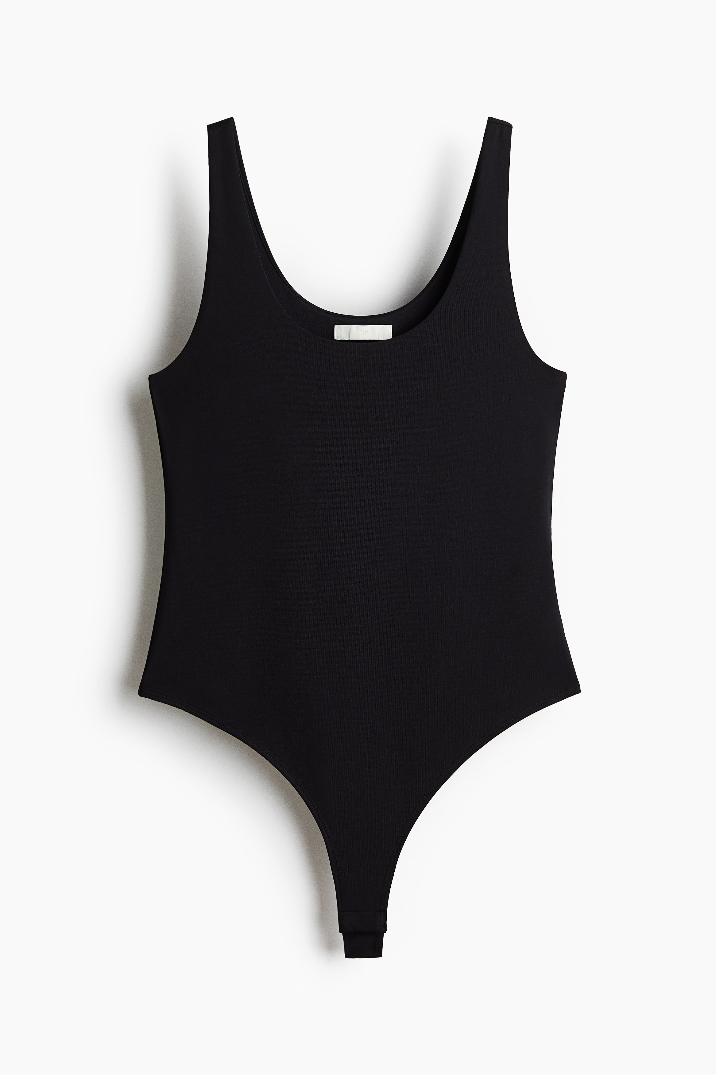 A black stretch-jersey bodysuit featuring a scoop neckline and wide shoulder straps is being worn by the model. The bodysuit is tucked into black jeans and styled with a cream-colored sweater draped over the shoulders.
