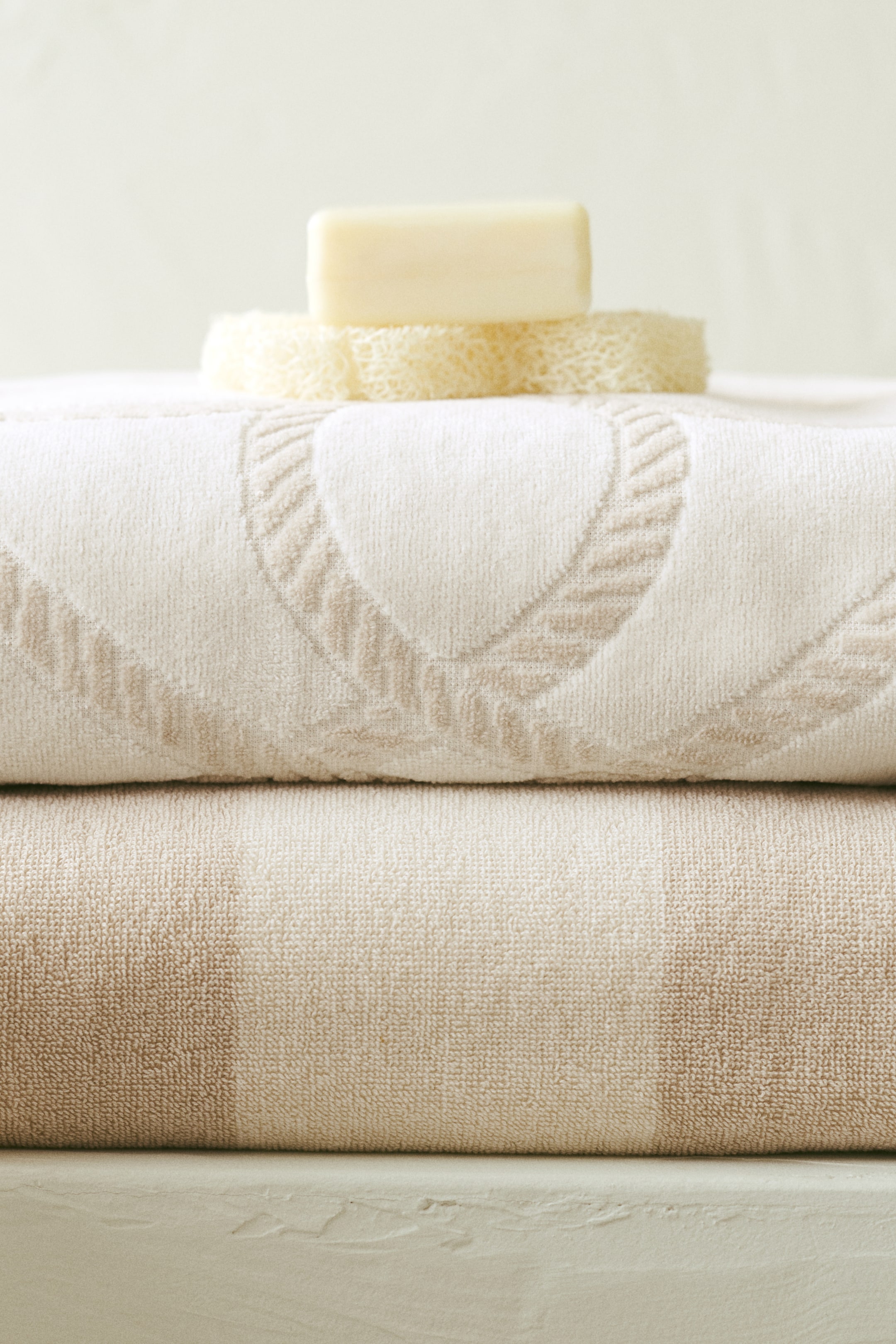 Visa större bild: En stapel vikta handdukar med en tvål och en loofah. Den översta benvita handduken har ett ljusbeige vävt mönster som liknar sammanflätade hjärtan, medan den nedre beige handduken visar subtila tonala ränder.