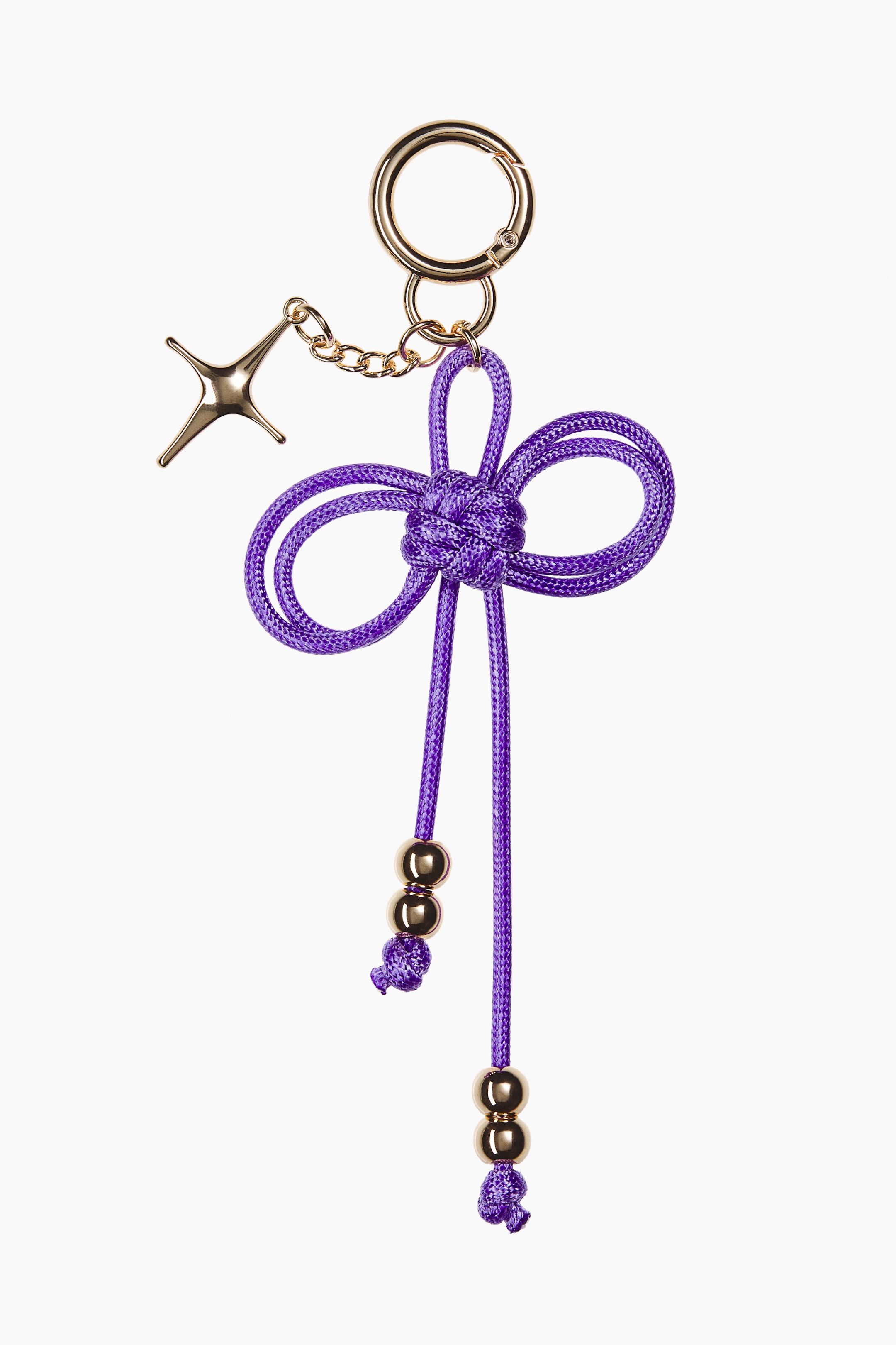 Ver imagen más grande: Un colgante para bolso presenta un diseño frontal con un cierre metálico en tono dorado, una cadena corta y un colgante metálico de cuatro puntas en forma de estrella. Un cordón morado está anudado de forma intrincada en un patrón decorativo similar a una flor, con dos cordones colgando en la parte inferior, cada uno adornado con dos cuentas esféricas en tono dorado.