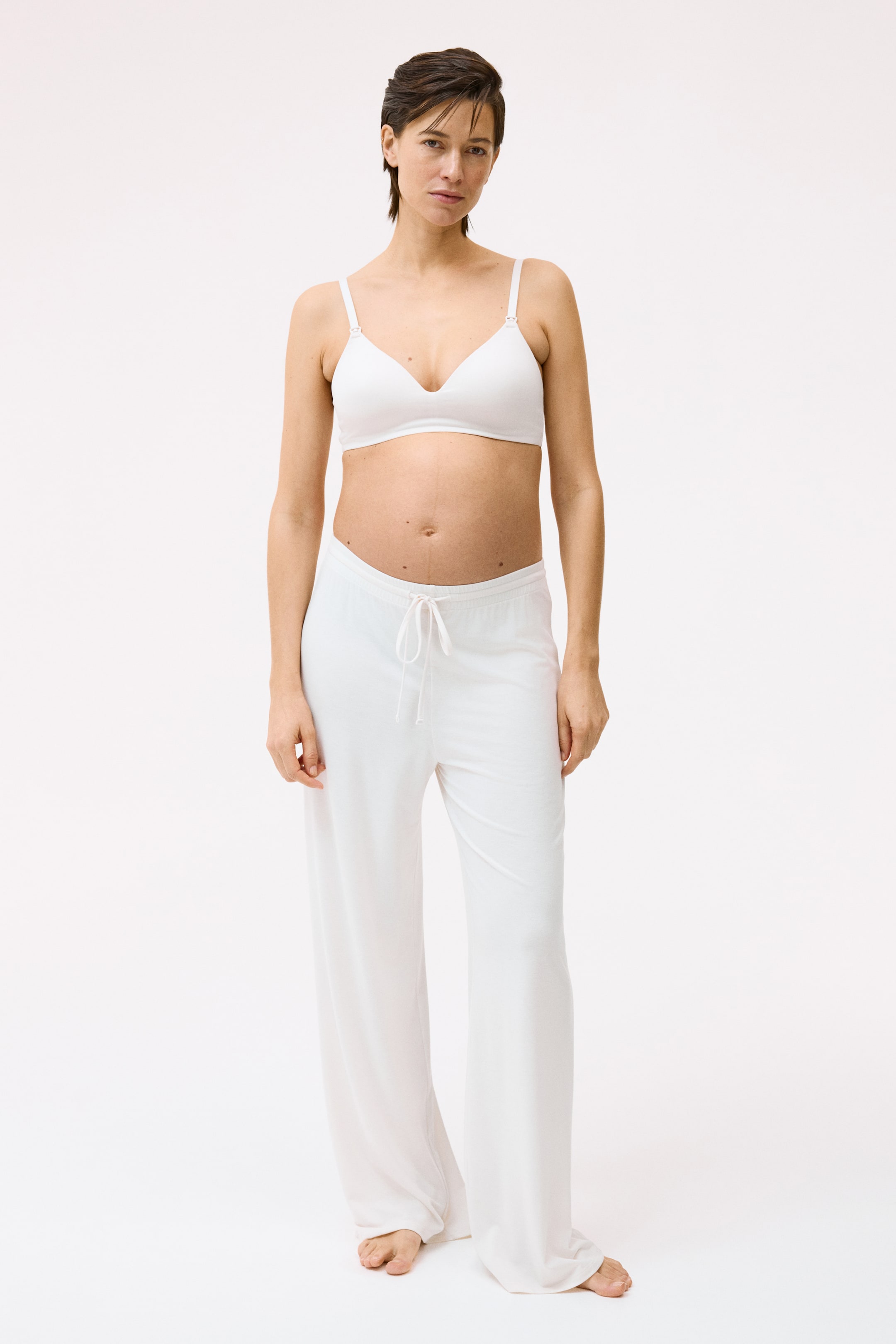 Se større billede: En hvid amme-bh og hvide loungebukser med brede ben vises forfra. Bh'en har tynde, justerbare stropper med synlige clips, og bukserne har en snøre i taljen.
