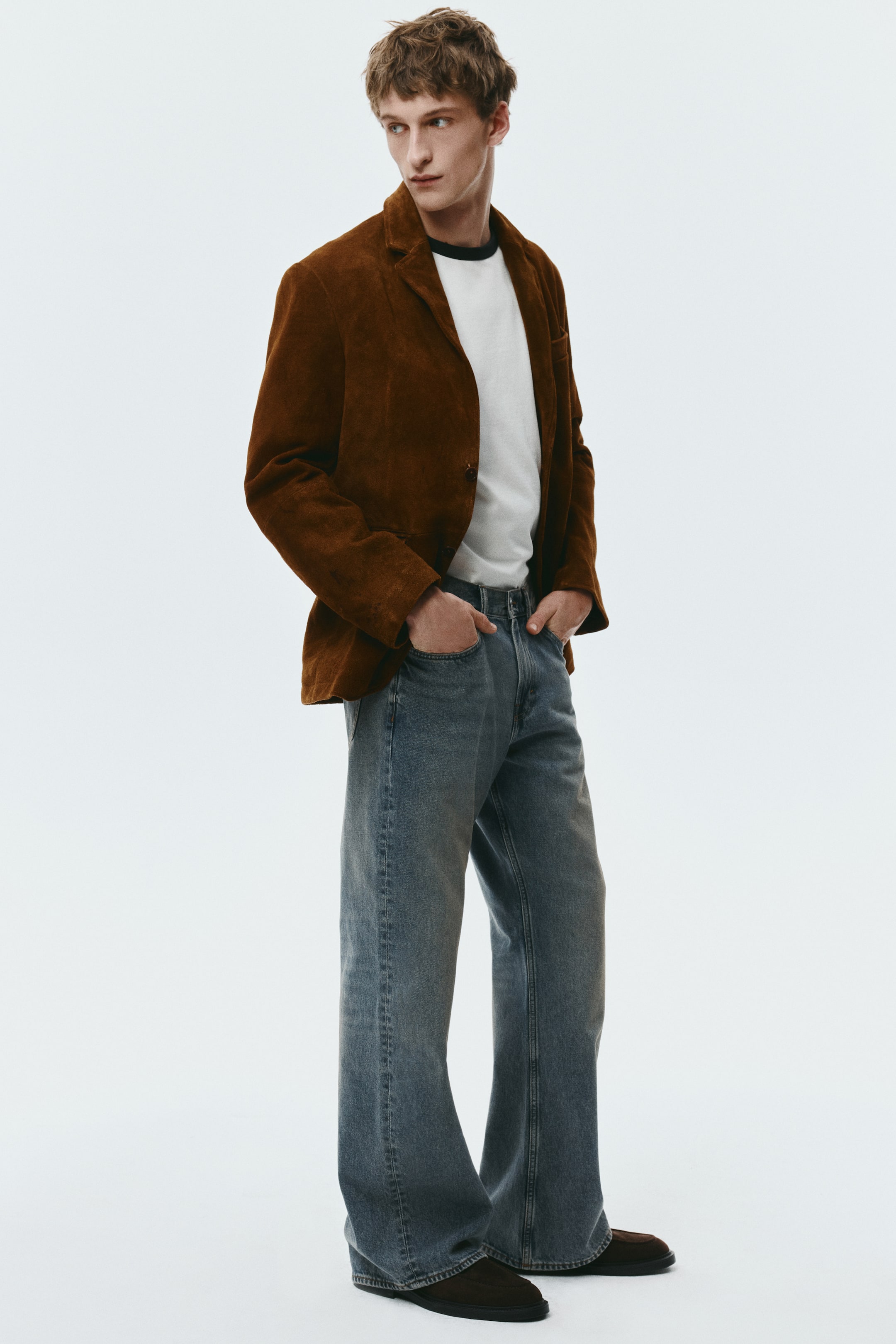 Agrandir l'image: Ce blazer en daim marron présente des revers crantés et une fermeture par un bouton apparent, présenté sous un léger angle trois-quarts. Il affiche une coupe décontractée et se porte en superposition sur un t-shirt blanc.