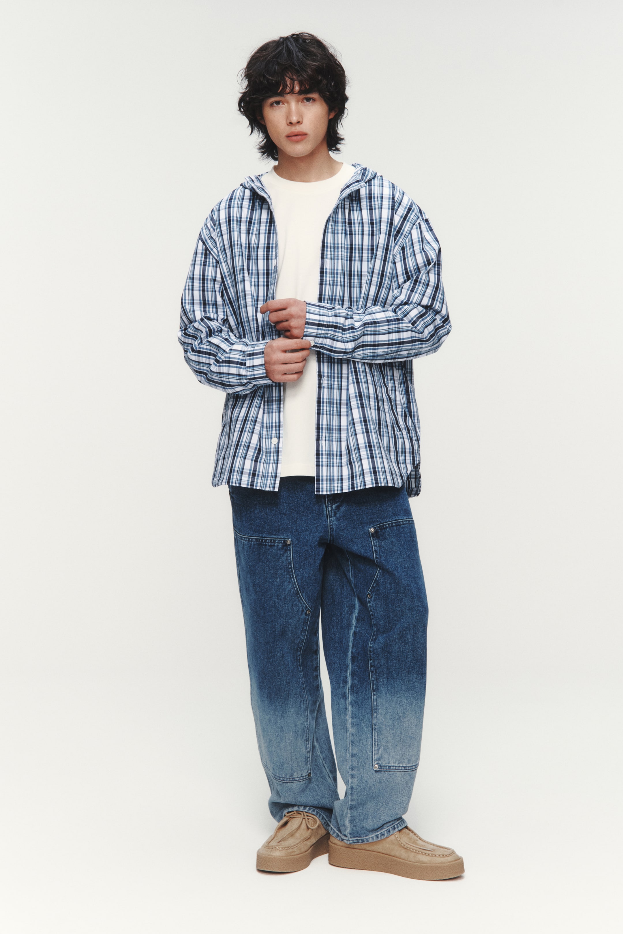 이미지 확대 보기: 모델이 입고 있는 옷은 파란색, 흰색, 검정색의 체크무늬 후드 셔츠로, 단추가 있는 앞부분이 열려 있으며 긴 소매에 단추가 있는 커프스가 있습니다. 모델은 느슨한 핏의 셔츠를 밝은 색상의 티셔츠 위에 레이어링하여 입고 있으며, 한쪽 커프스를 조정하고 있습니다.