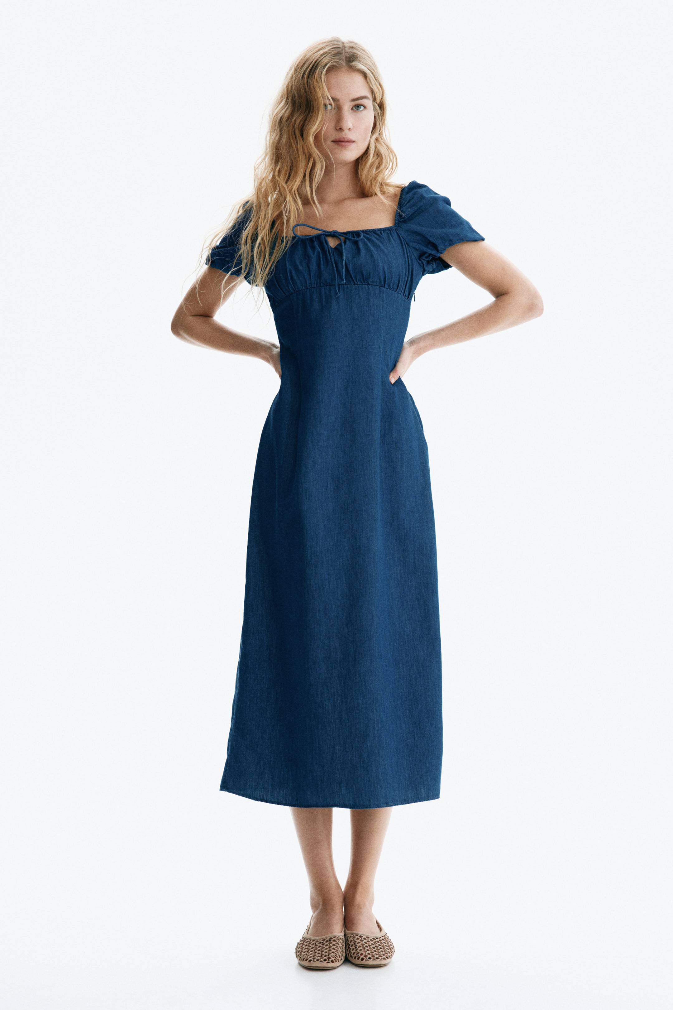 View larger image: A dark blue denim midi dress featuring a square neckline, puffed short sleeves and a ruched bodice with a front-tie detail, fitted at the waist and flowing into an A-line skirt.
