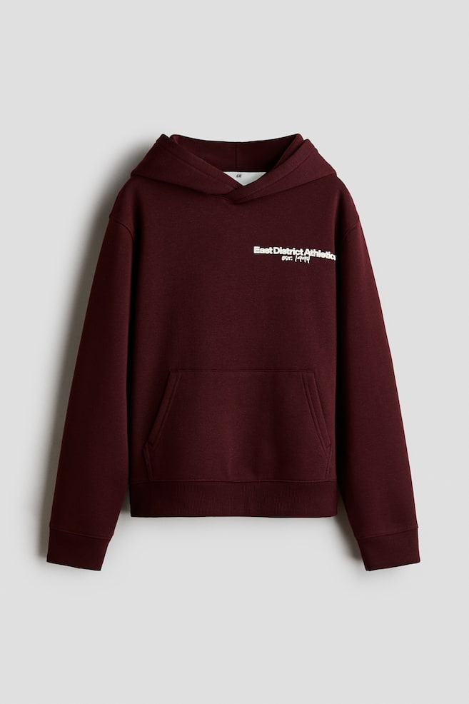 Ein dunkelburgunderfarbener Hoodie ist von vorne zu sehen, mit einer gefütterten Kapuze mit Wickel-Detail, überschnittenen Schultern und einer Kängurutasche. Auf der linken Brust befindet sich ein kleiner weiÃer Schriftzug mit der Aufschrift âEast District Athletic EST. 1994â. Abgerundet wird der Hoodie durch Rippbündchen an Ãrmeln und Saum. - 1