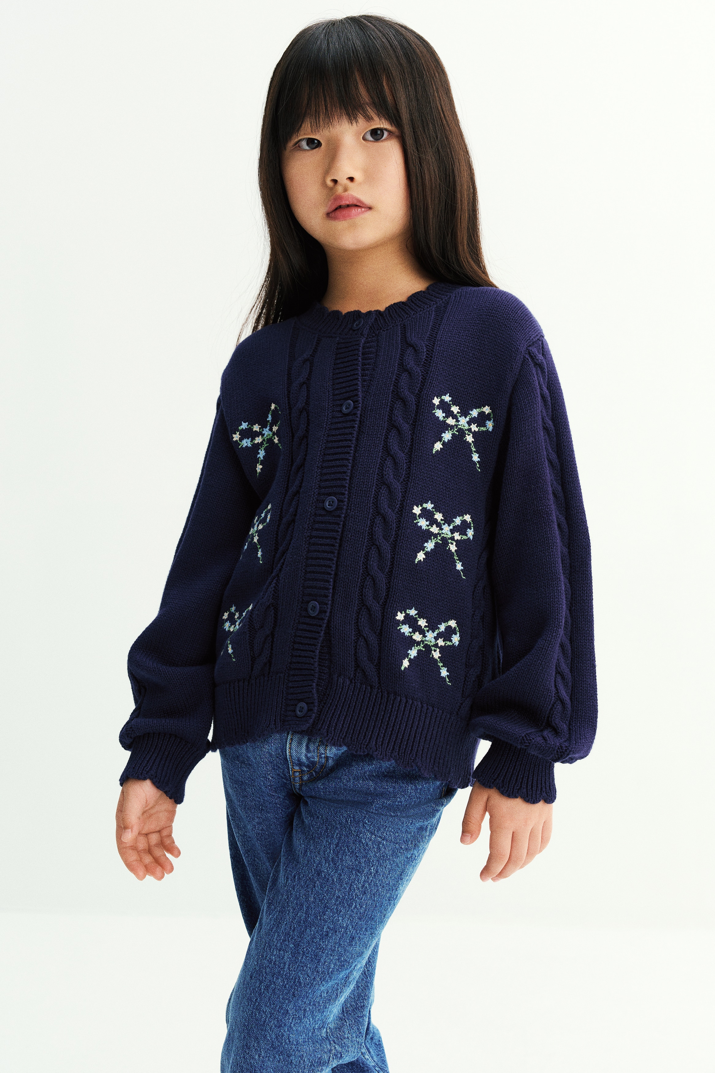 A navy blue knit cardigan is shown from the front, featuring a round neckline, button-down closure, and cable-knit details along the front and sleeves. Six embroidered floral bow motifs decorate the chest area, with scalloped edges finishing the ribbed collar, cuffs, and hem.