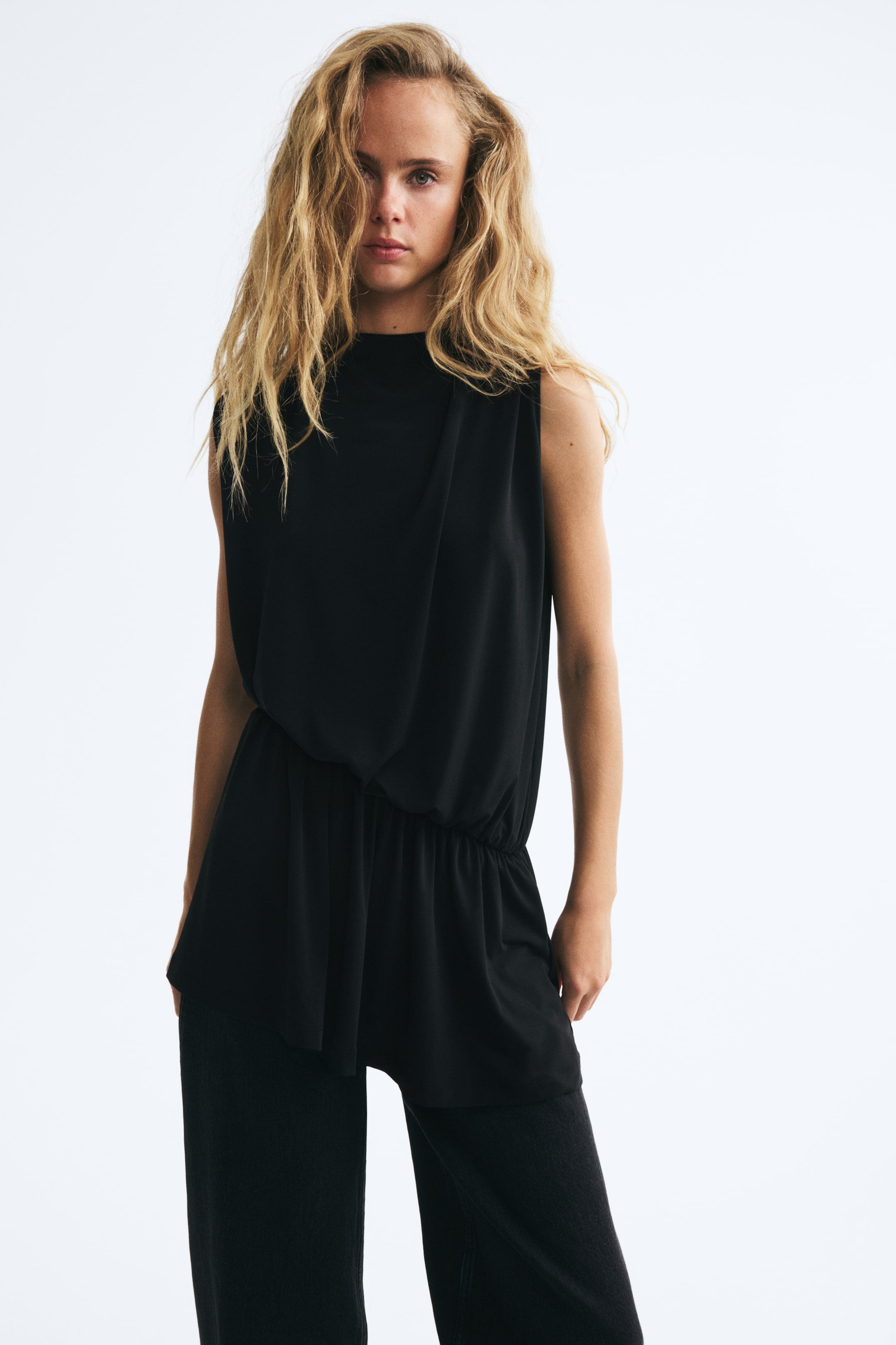 View larger image: A loose-fit draped top in black is worn by the model, featuring a high neckline, sleeveless design, and a draped front that gathers into an asymmetrical waist. The top is styled with coordinating black trousers.