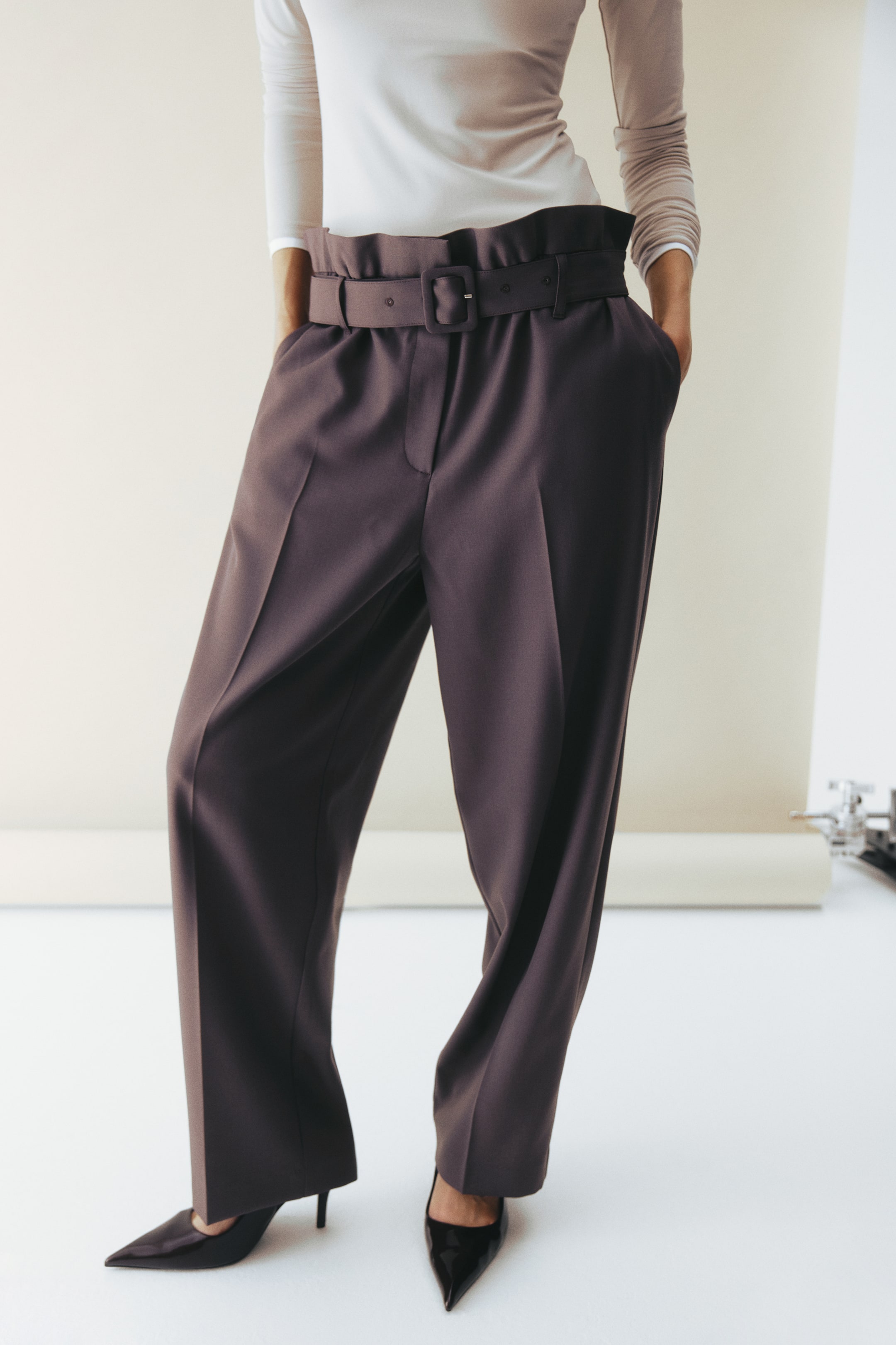 View larger image: Deep plum-brown wide-leg pants feature a high paperbag waist with ruffles and a matching wide belt with a rectangular buckle. The pants present a relaxed fit with front pleats, side pockets and a full-length cut, styled with a tucked-in top and black pointed-toe shoes.