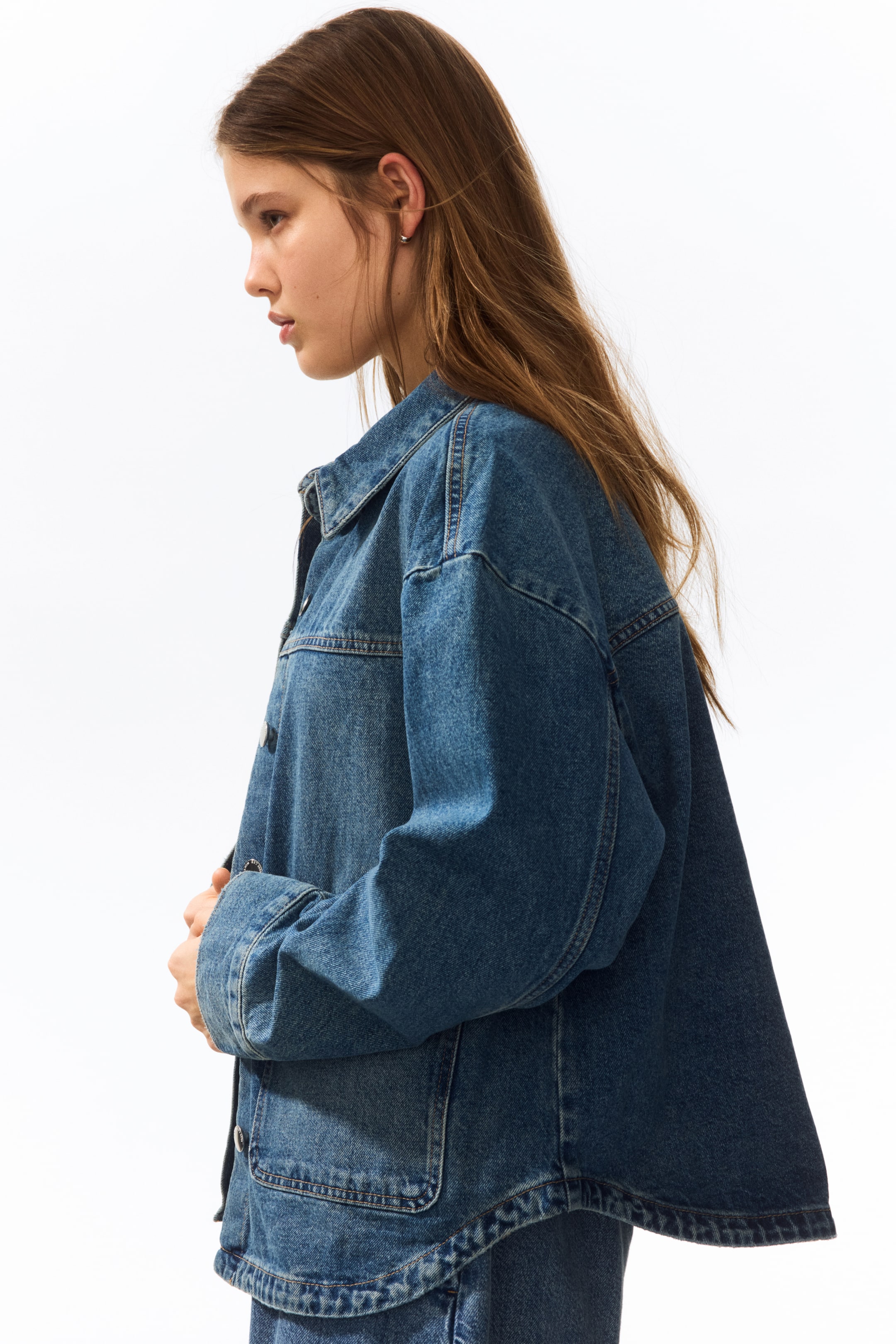 View larger image: A medium blue denim jacket is being showcased from a side angle, highlighting its oversized fit and dropped shoulders. It features a pointed collar, visible front buttons, and a large patch pocket on the lower front, with contrasting stitching.