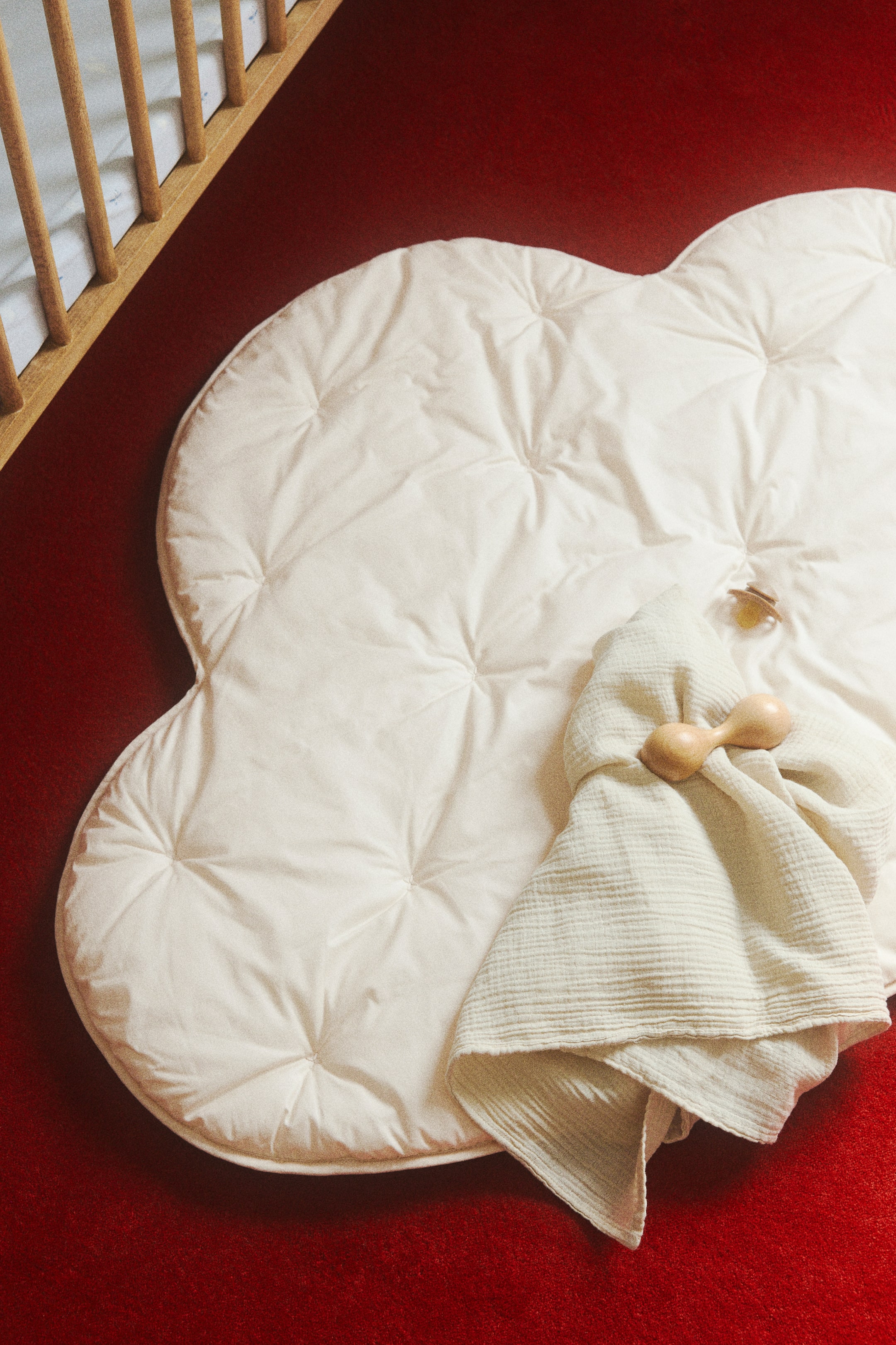 View larger image: A cream-colored, cloud-shaped mat lies flat, showcasing a tufted design that forms puffy sections. A light-colored, textured blanket is neatly folded on the mat, accompanied by a wooden rattle and a pacifier resting on its surface.
