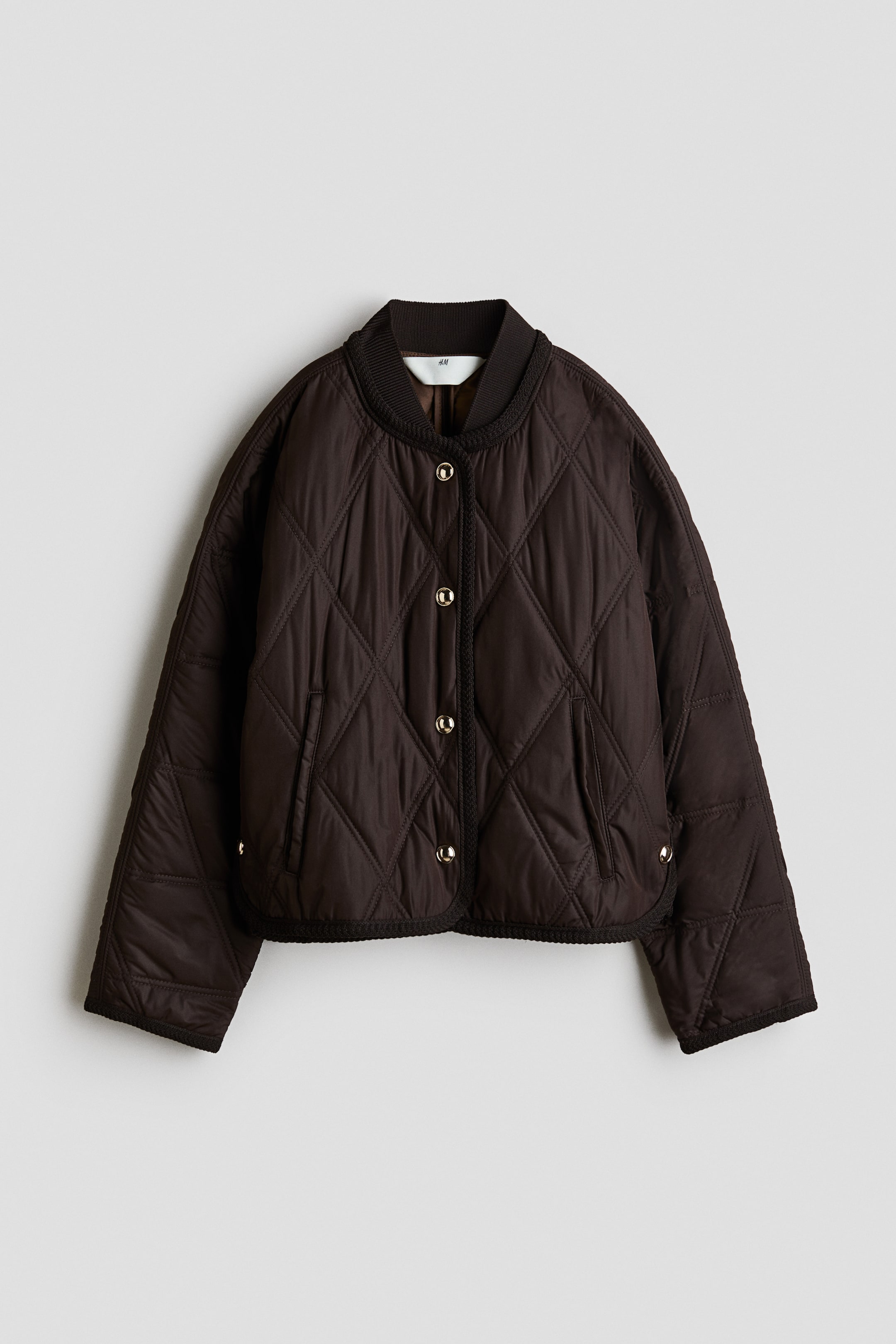 Grotere afbeelding bekijken: VOORKANT, een donkerbruine doorgestikte bomberjas toont een ruitpatroon. Hij heeft een geribde opstaande kraag, vijf goudkleurige drukknopen middenvoor en twee schuine paspelzakken. Een gevlochten zwarte bies accentueert de sluitlijst, kraag, zoom en manchetten.