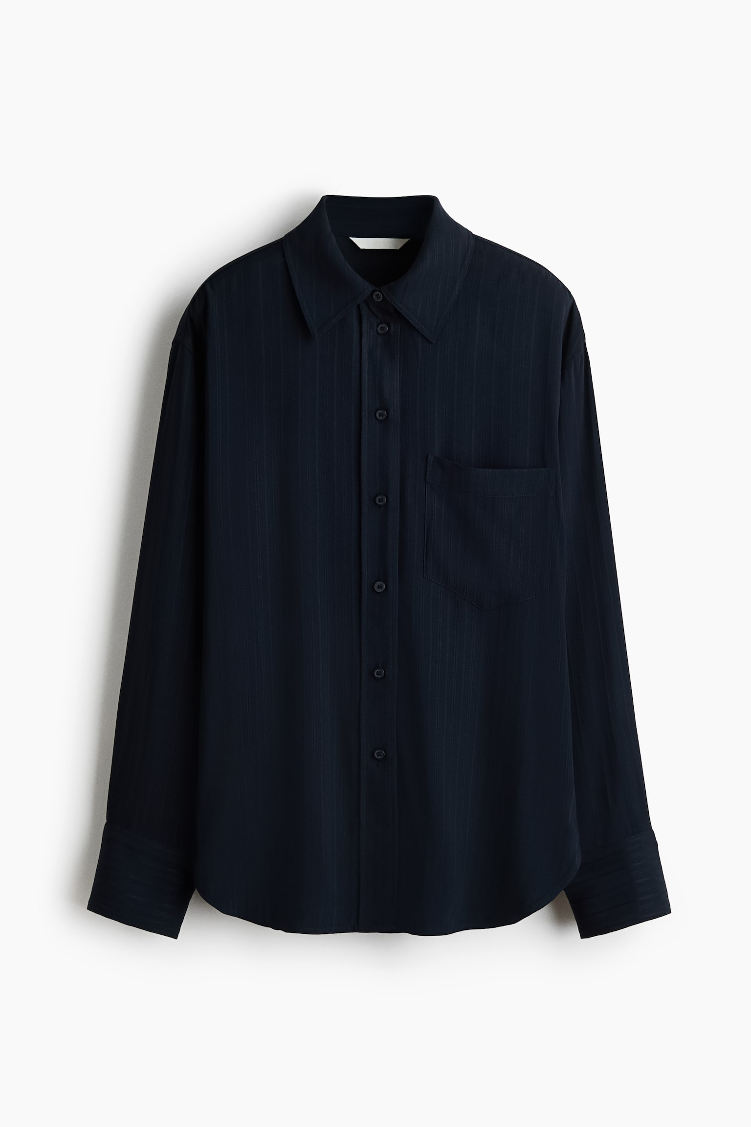 Una camicia oversize blu navy scuro Ã¨ appesa frontalmente, mettendo in risalto una delicata trama testurizzata, un colletto a punta e una chiusura frontale con bottoni. Il design presenta una singola tasca applicata sul petto, spalle scese, maniche lunghe con polsini larghi e un orlo arrotondato.