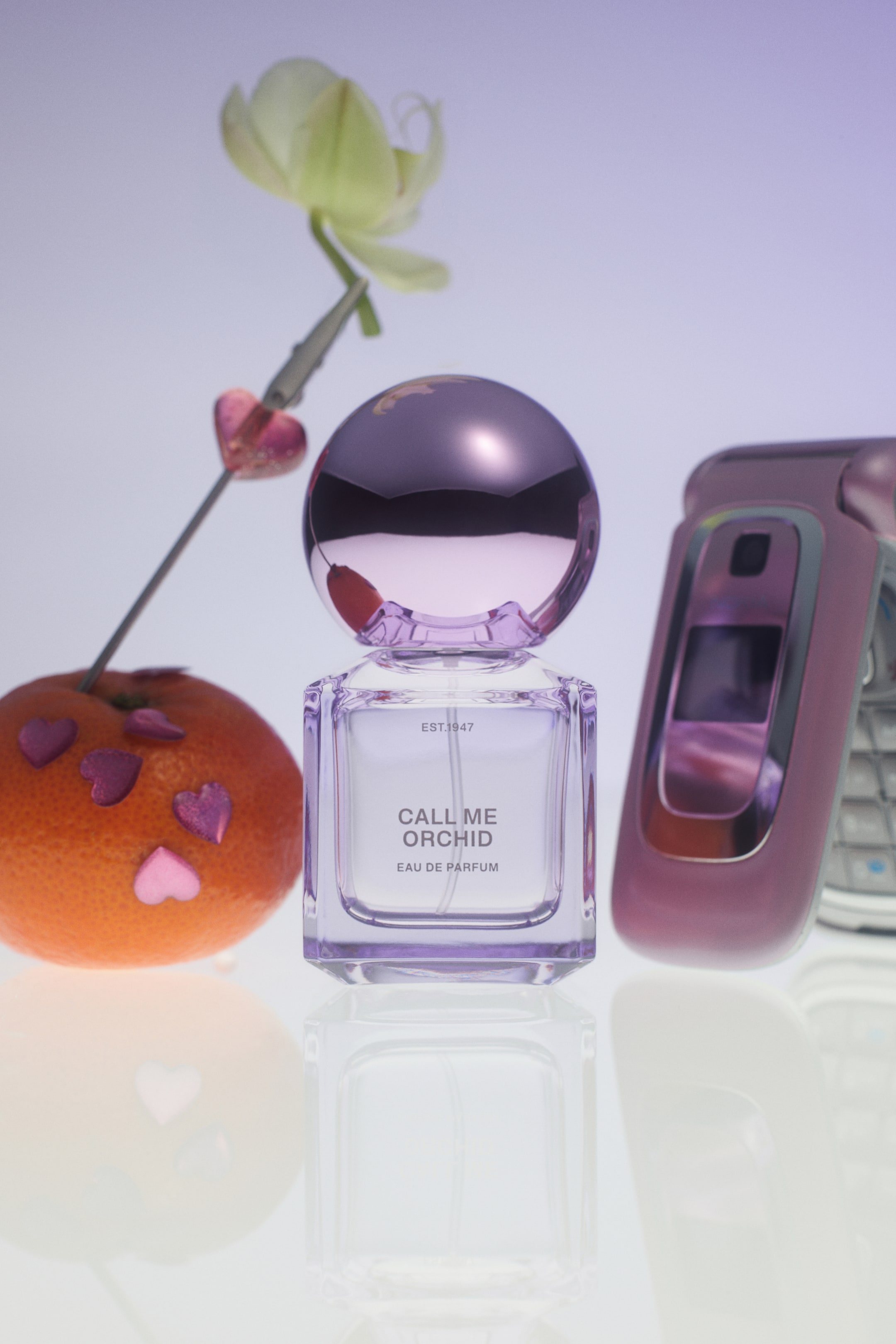 View larger image: A square glass bottle of "Call Me Orchid" Eau de Parfum stands upright on a reflective surface, featuring a light purple tint and a shiny metallic spherical cap in a matching purple shade. The bottle is flanked by a mandarin orange decorated with small pink heart stickers on the left and a pink flip phone on the right.