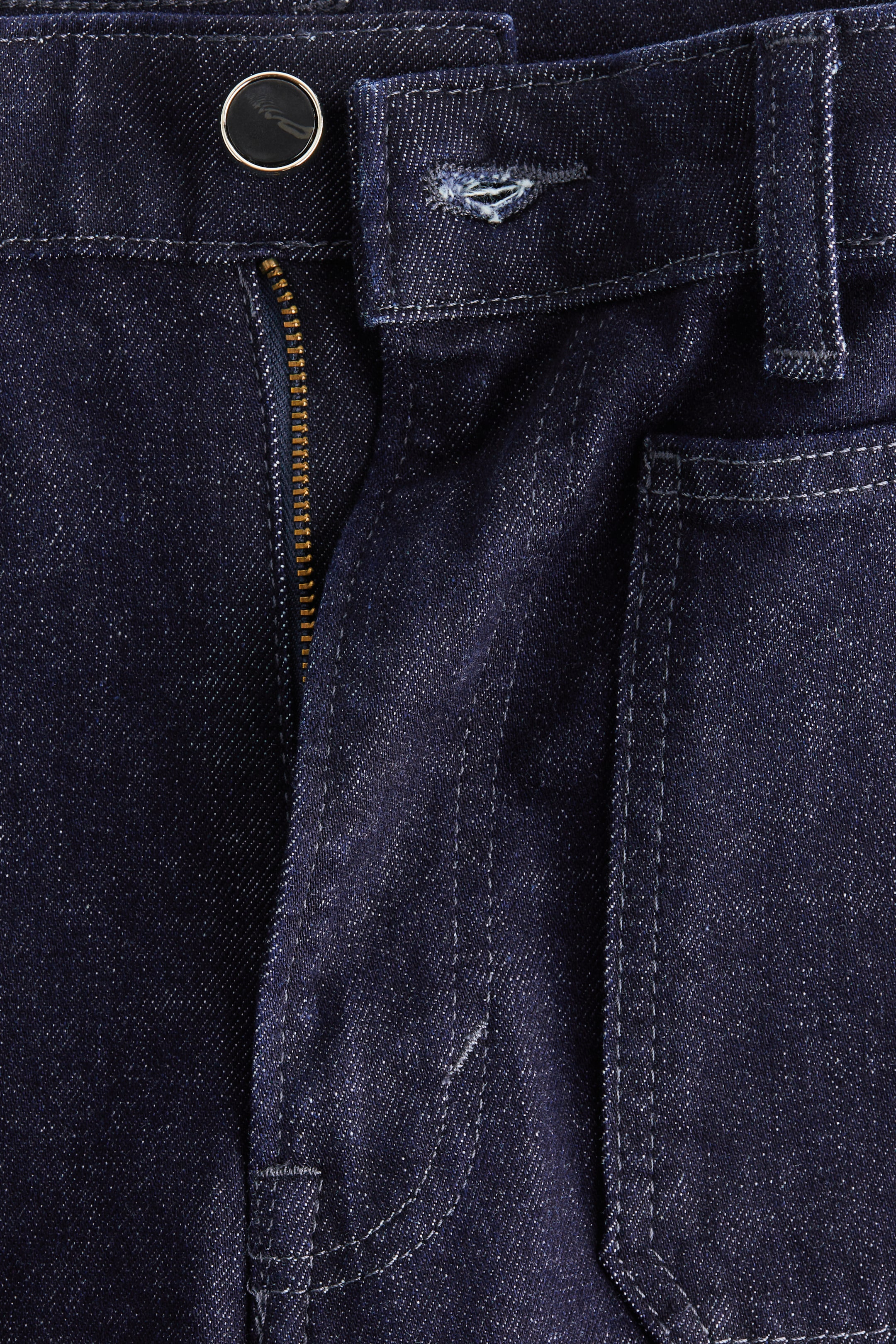 Se større billede: Forsiden af mørkeblå denimjeans har et bælte fastgjort med en sort knap med en metallisk kant. En messingfarvet lynlås er delvist synlig på gylpen, og lysfarvede stikninger fremhæver bæltet og en forreste lomme.