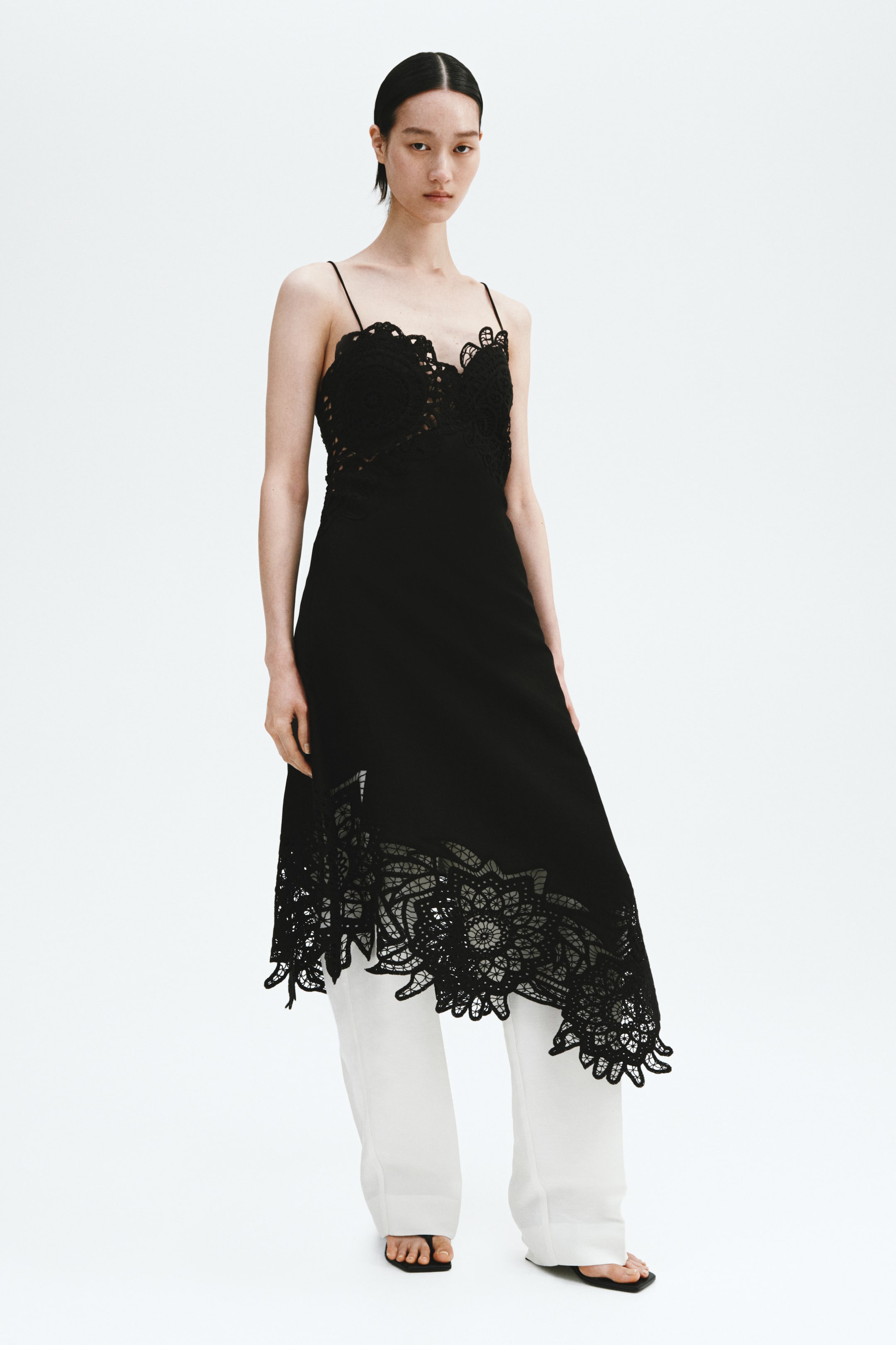 Ingrandisci l'immagine: Un abito nero frontale presenta delicati spalline a spaghetti e un intricato corpetto in stile bustier all'uncinetto, mentre la sua gonna fluida mette in mostra un orlo con dettagli in pizzo a ritaglio su larga scala, stratificato su pantaloni bianchi.