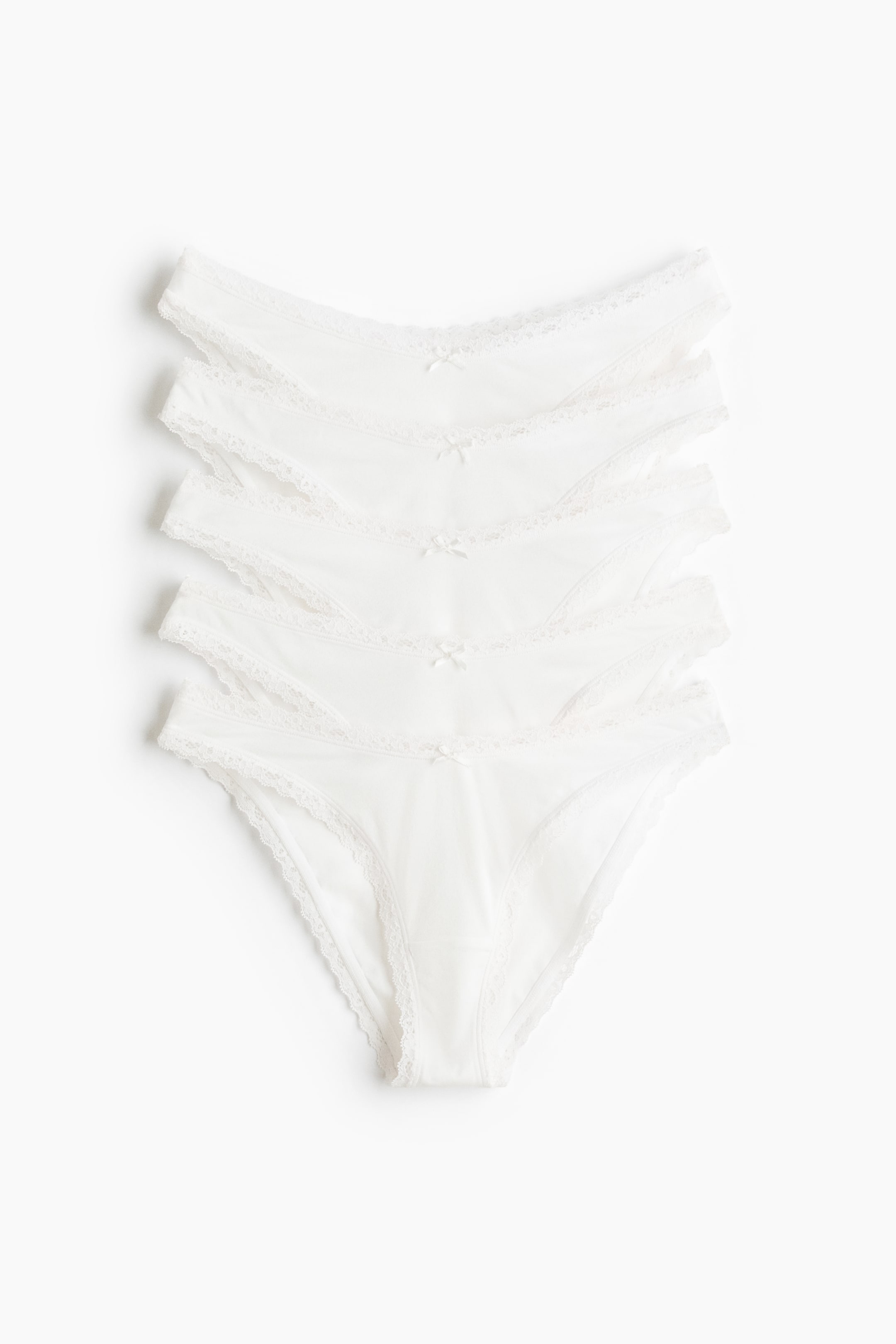 Grösseres Bild ansehen: Baumwoll-Jersey-Slips sind ausgelegt und zeigen ihre Vorderansicht mit einer tiefen, V-förmigen Taille. Jeder weiße Slip ist mit zarten Spitzendetails entlang der Taille und Beinausschnitte verziert und hat eine kleine dekorative Schleife an der Vorderseite.