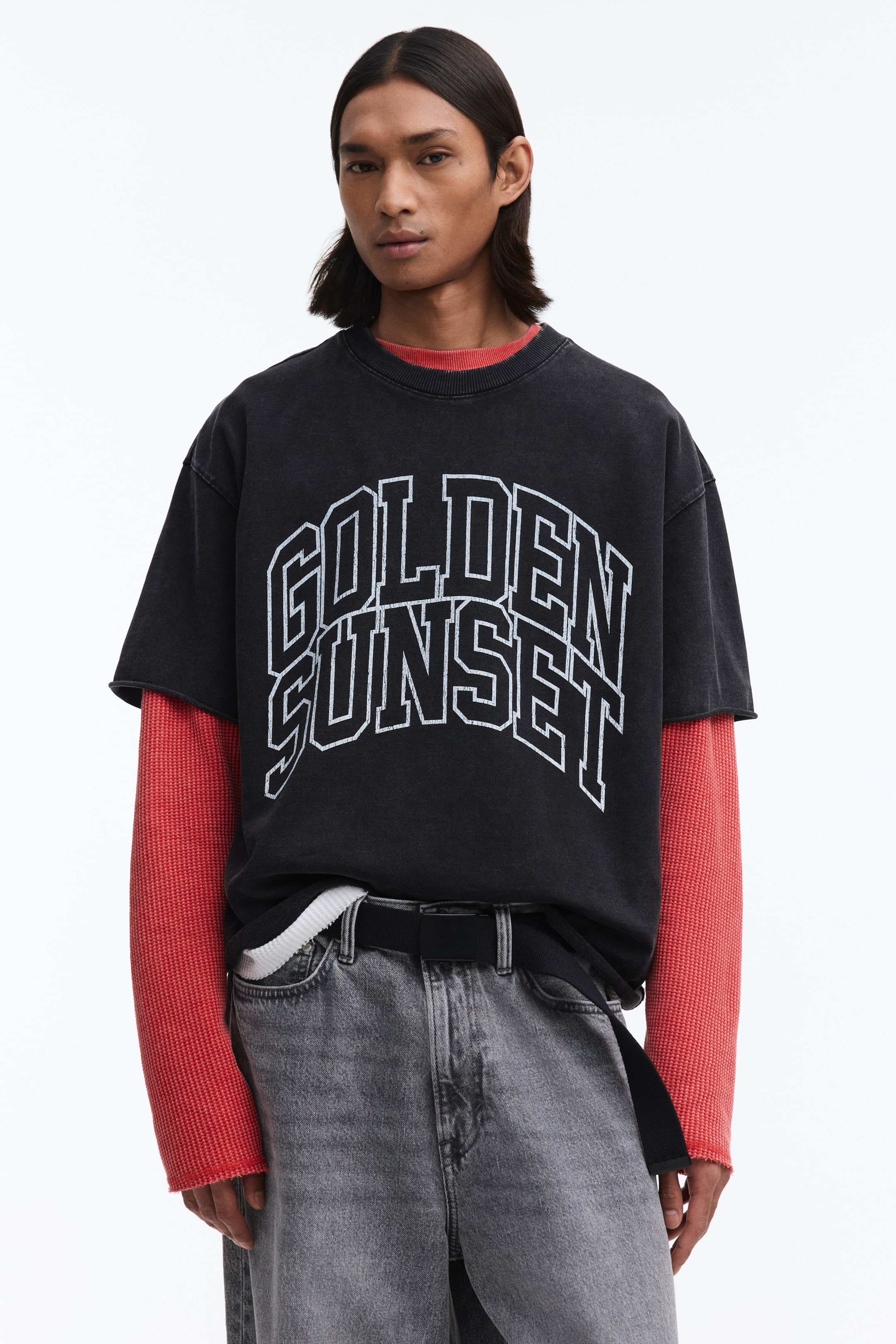 Ampliar la imagen: Una camiseta negra de efecto lavado con mangas cortas, que presenta el texto "GOLDEN SUNSET" en blanco desgastado en el pecho, se combina con una camiseta roja de manga larga de tejido waffle, mostrando su cuello redondo y mangas con borde sin rematar.