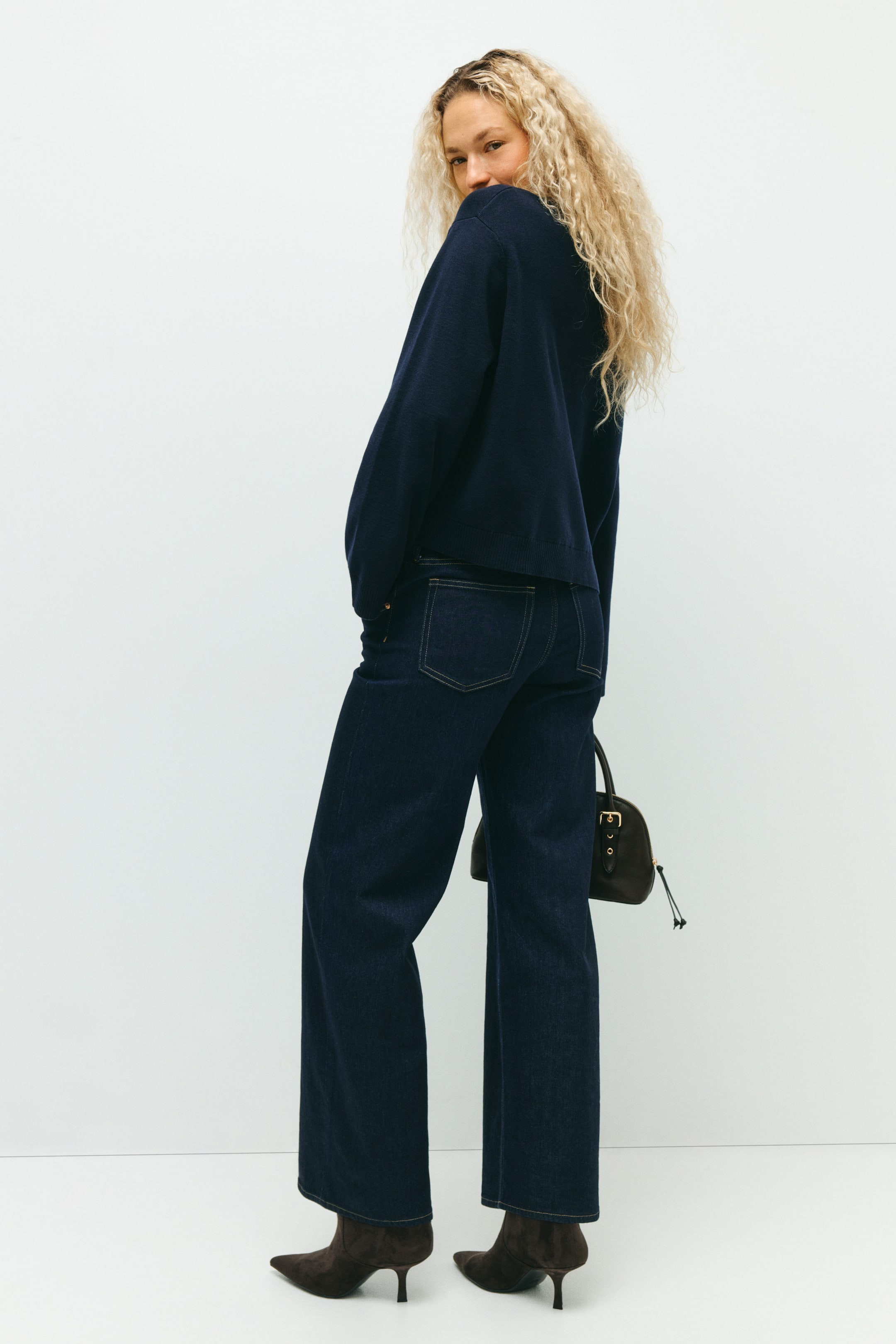 View larger image: A relaxed-fit navy knit sweater, featuring ribbed cuffs and hem, is being shown from the back and right side, paired with dark wash wide-leg jeans. The jeans feature contrast-stitched back pockets and are styled with a small dark brown handbag and pointed-toe heeled ankle boots.