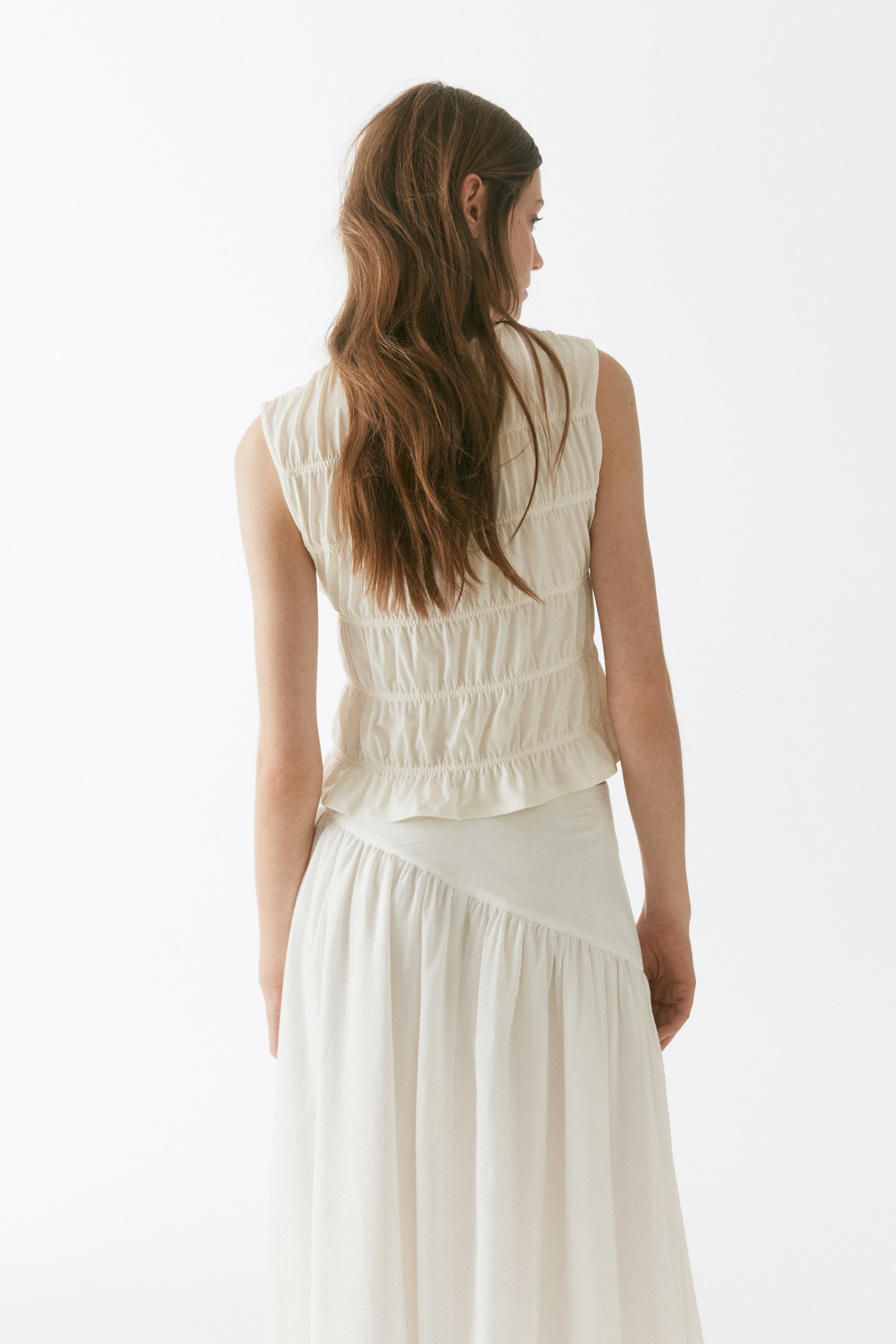 View larger image: The model is wearing a sleeveless smocked top crafted from stretch jersey, shown from the back. This top showcases smocking detail across the back, a keyhole opening with a button closure at the back of the neck, and a frill trim at the hem. It is paired with a matching light-coloured skirt.
